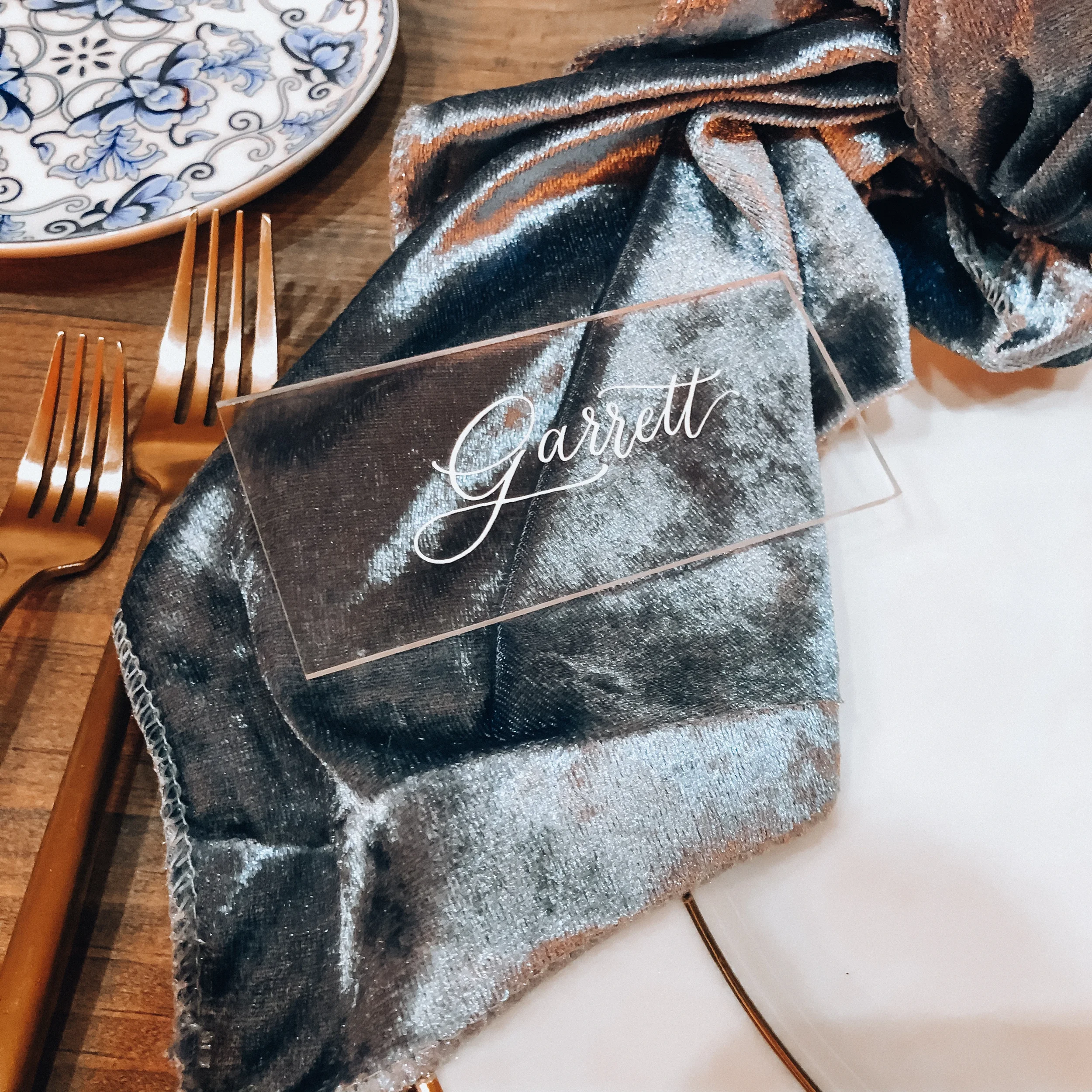 Perfect for a romantic, texture-driven reception table, this setting pairs a clear acrylic place card with ethereal white script calligraphy. Layered gracefully over a plush slate-blue velvet napkin and paired with vintage copper flatware, it creates