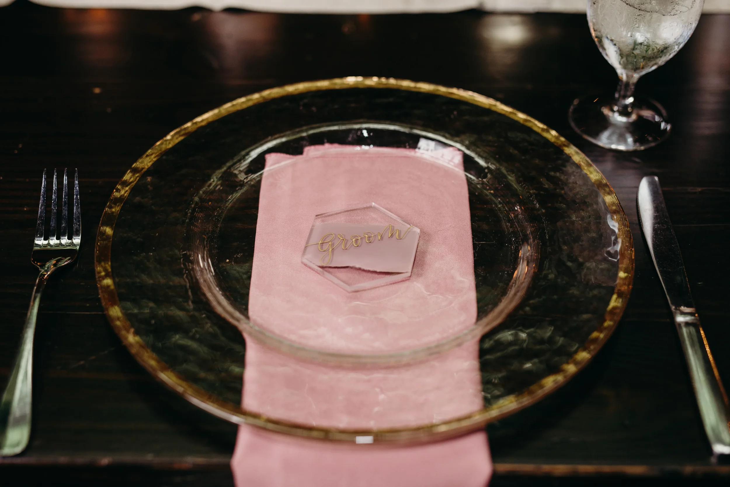 Elevate your fine-art tablescape with custom geometric clear acrylic place cards. Featuring fluid gold script calligraphy, this hexagon name card is layered over a dusty pink napkin and a textured glass charger plate to create a deeply romantic, high