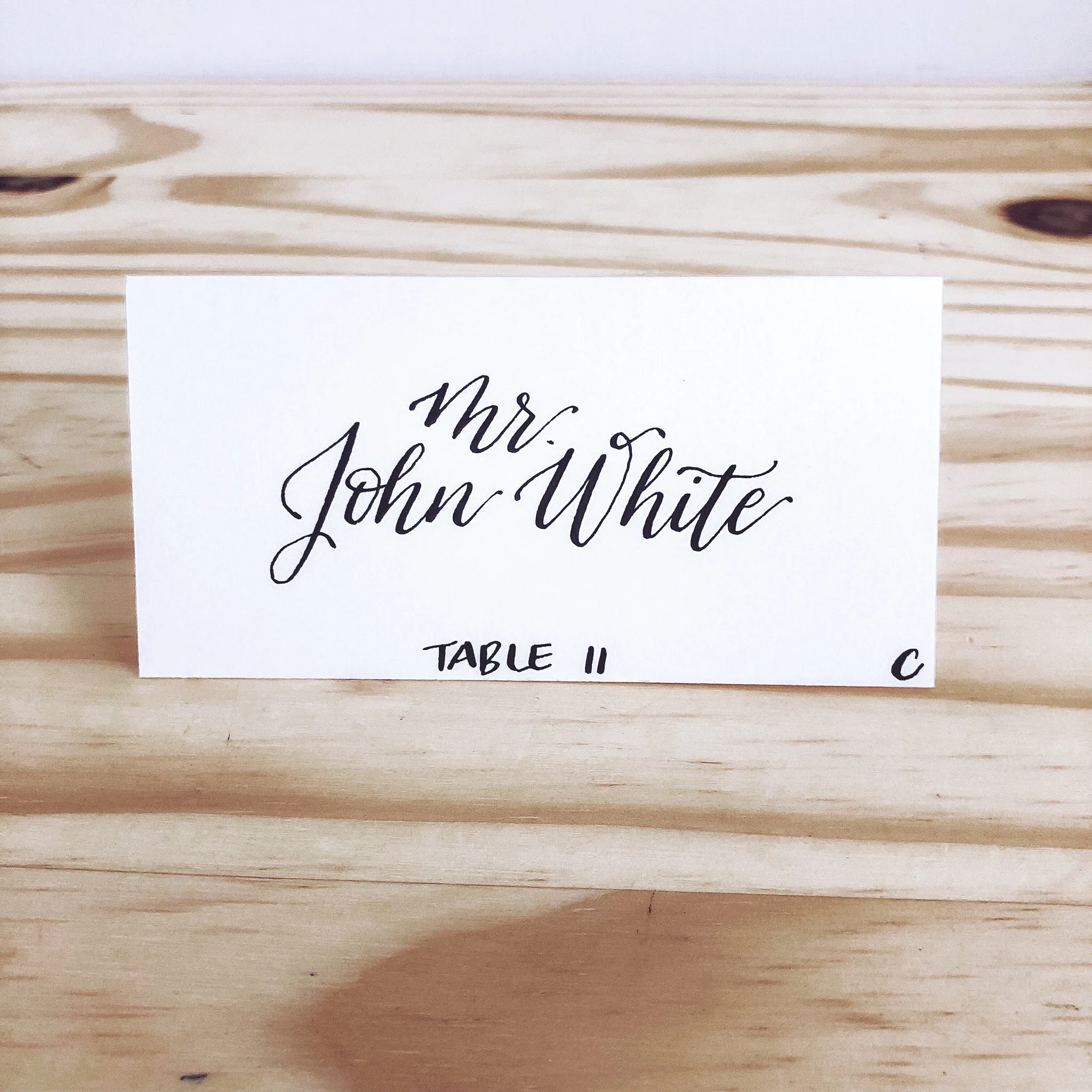 Minimalist white folded wedding place card with sharp black calligraphy script reading "Mr. John White."