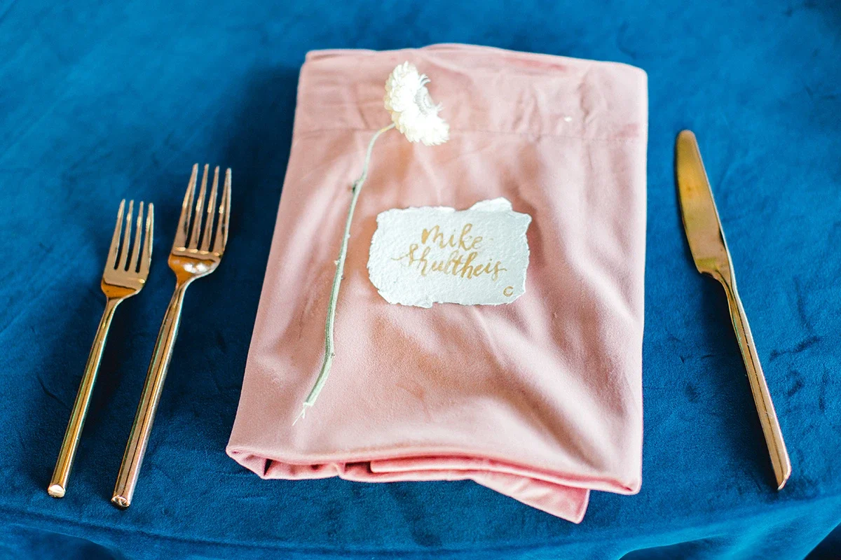 High-angle view of fine-art wedding place setting with gold ink guest card, modern gold flatware, and peach linen.