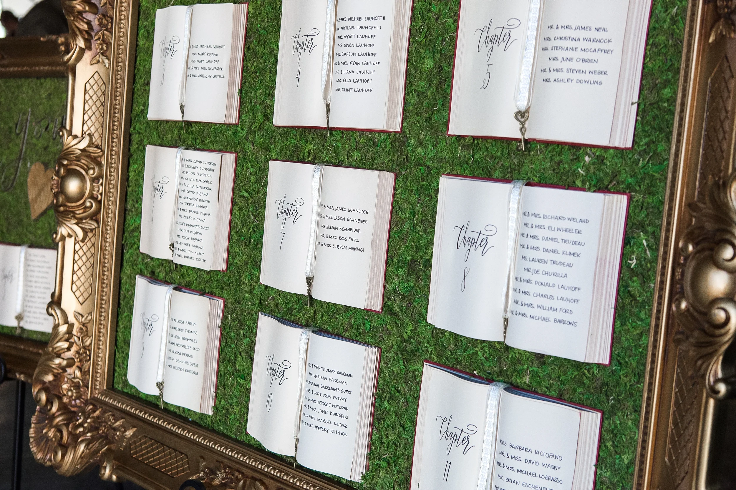 Storybook-themed wedding seating chart with vintage open hardcover books hand-lettered with calligraphy on a preserved moss backdrop.