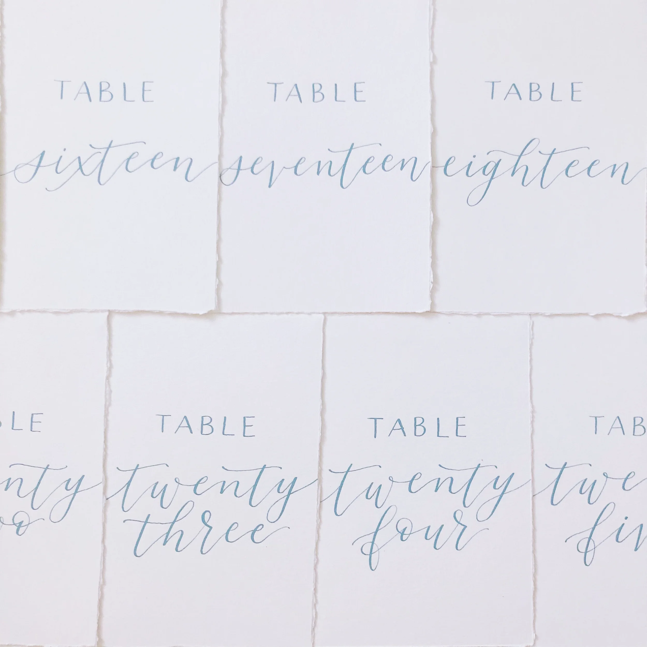 Deckled-edge handmade paper cards displaying written table numbers in dusty blue calligraphy script.