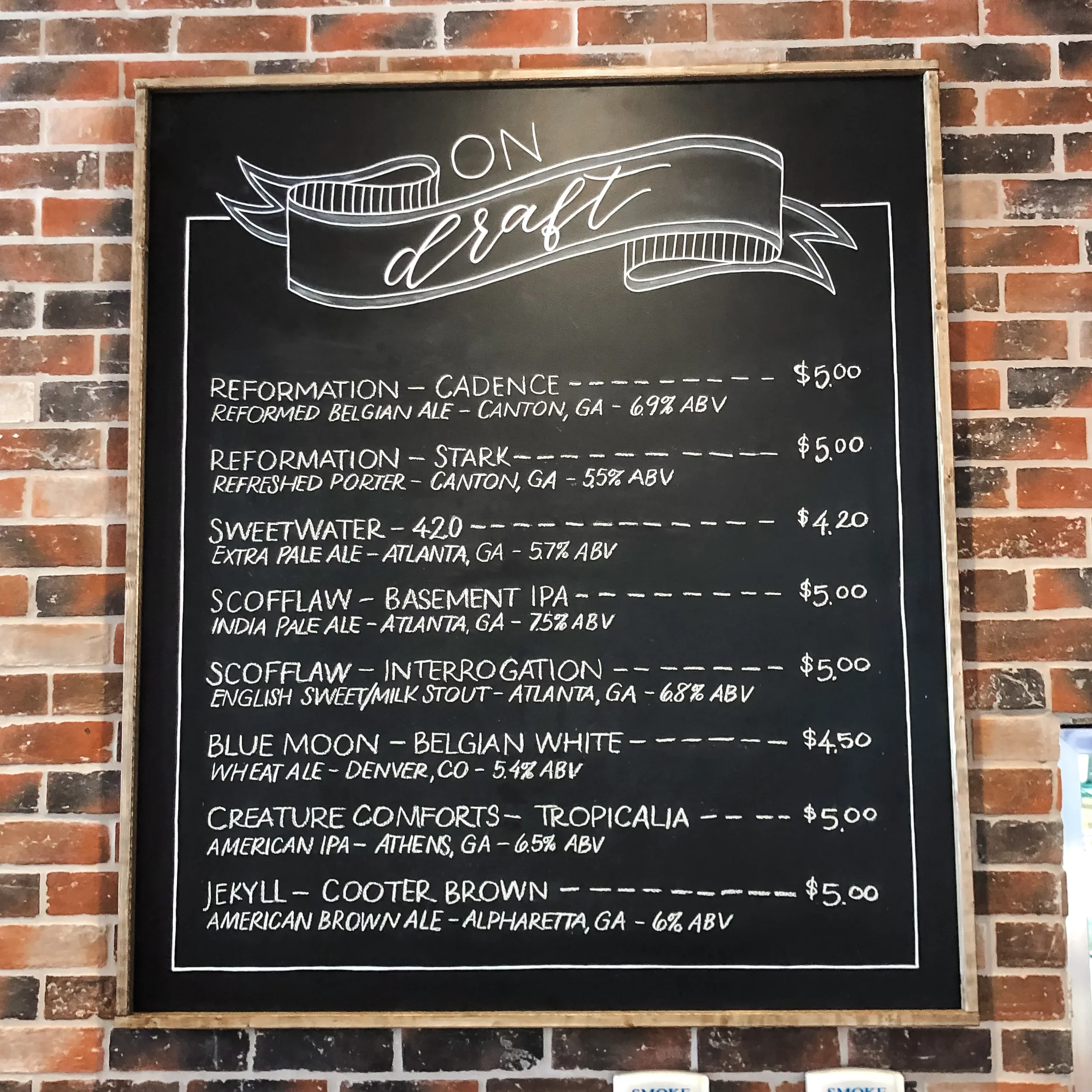 Large framed chalkboard bar menu titled "On Draft" hanging on a rustic red brick wall.