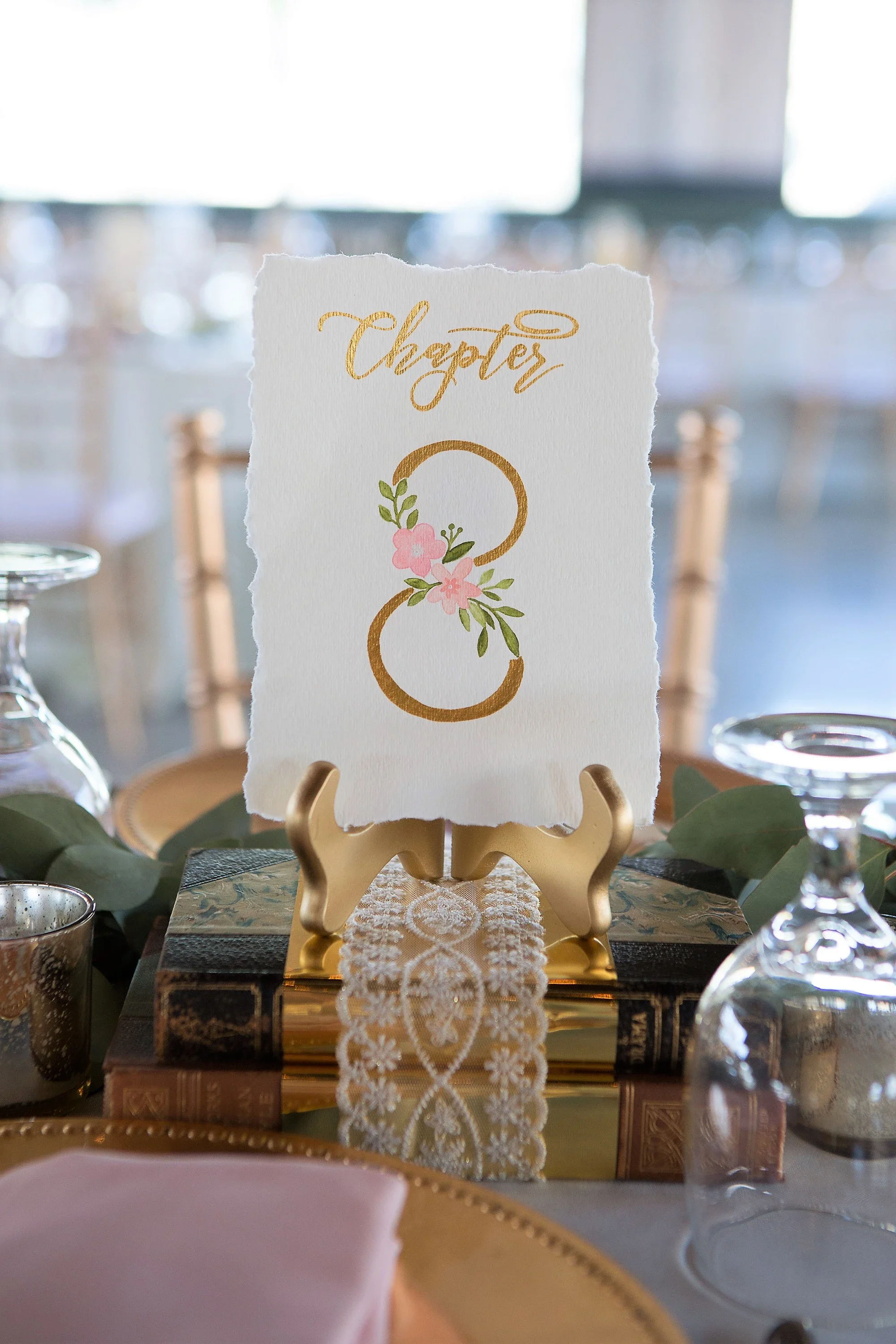 Deckled-edge table number reading "Chapter 8" with gold script and painted pink flowers, displayed on vintage books.