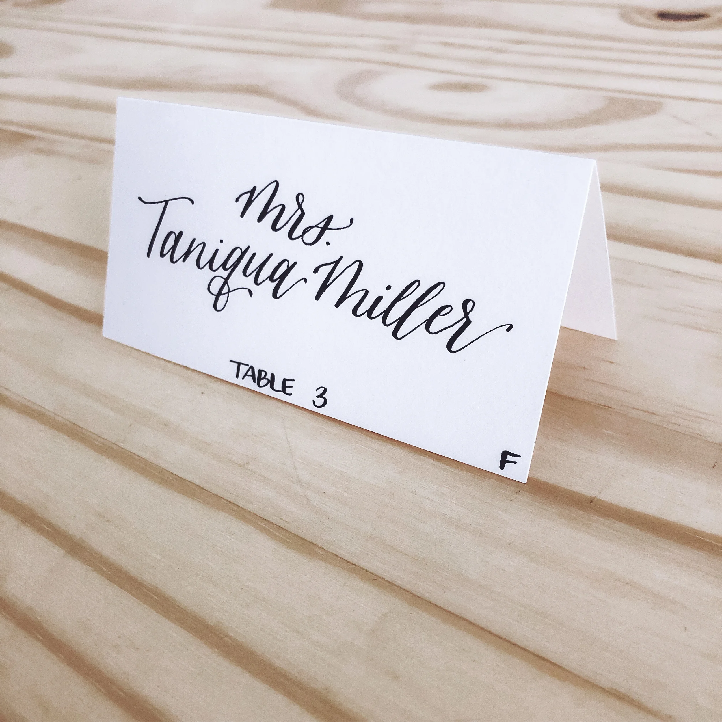 Minimalist white folded wedding escort card with sharp black calligraphy script.