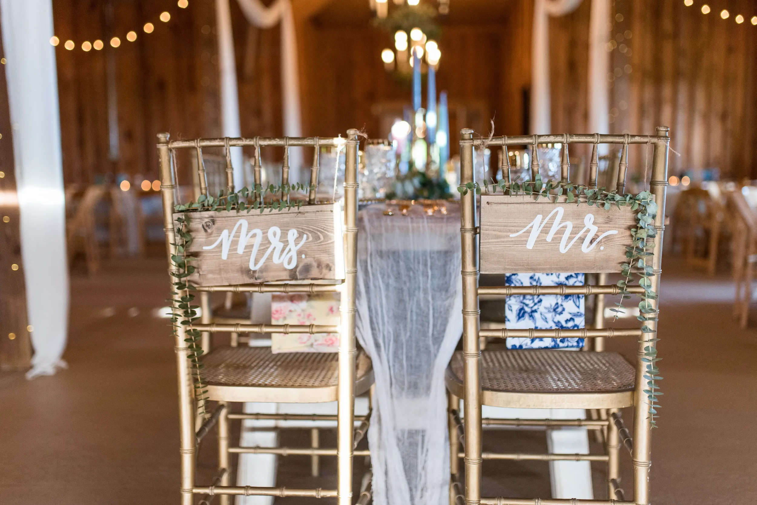 Make your sweetheart table unforgettable with personalized wooden wedding chair signs. Adorned with delicate greenery runners, these custom hand-lettered pieces add a luxurious, fine-art twist to a rustic barn reception setting.