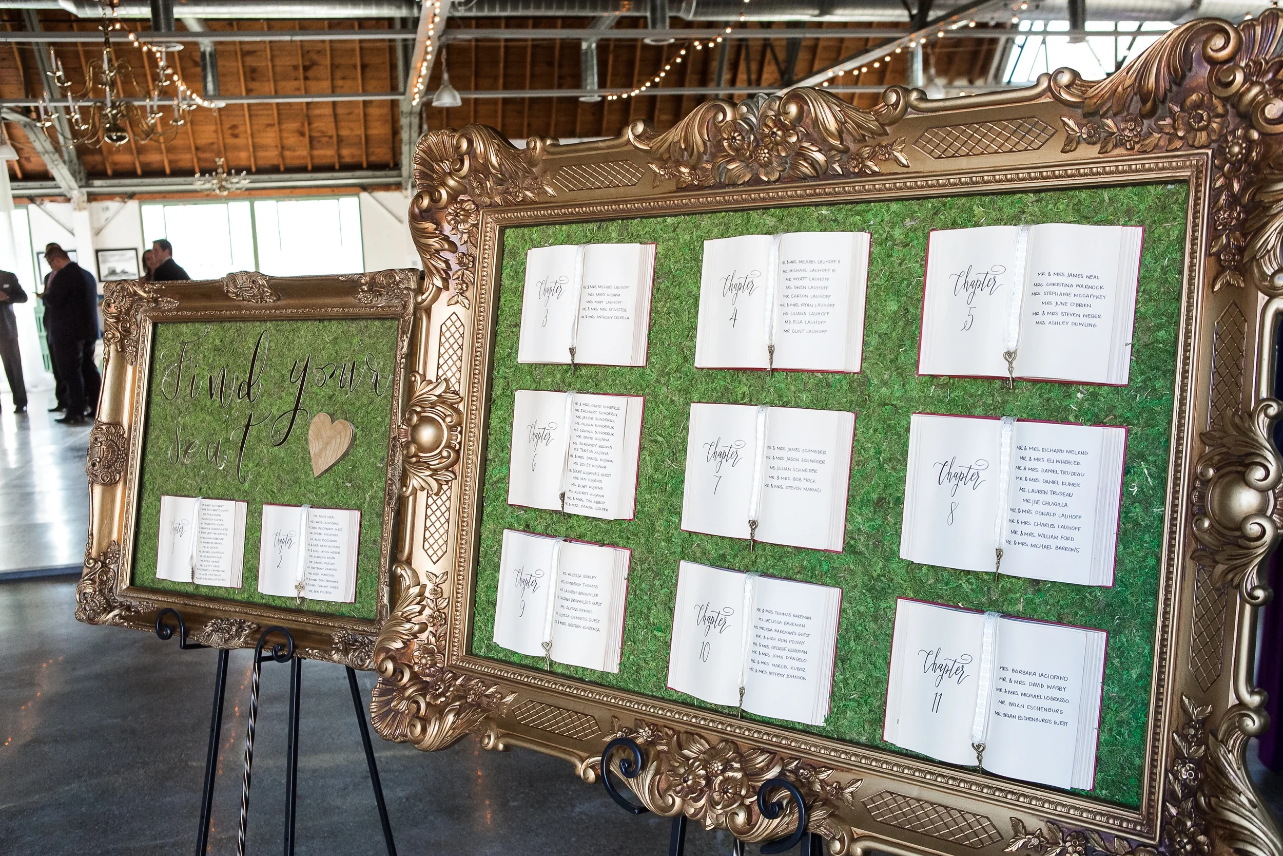 Ornate gold baroque frame mounted over a green moss wall featuring open books lettered with "Chapter" table numbers and guest lists, accented by key bookmarks.