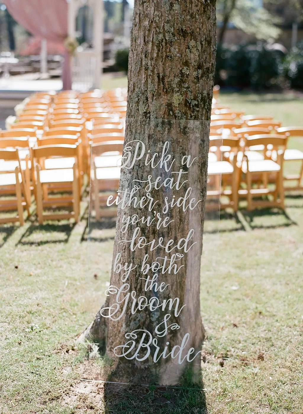 Clear acrylic custom wedding ceremony quote sign that says "pick a seat not a side" in white script calligraphy.
