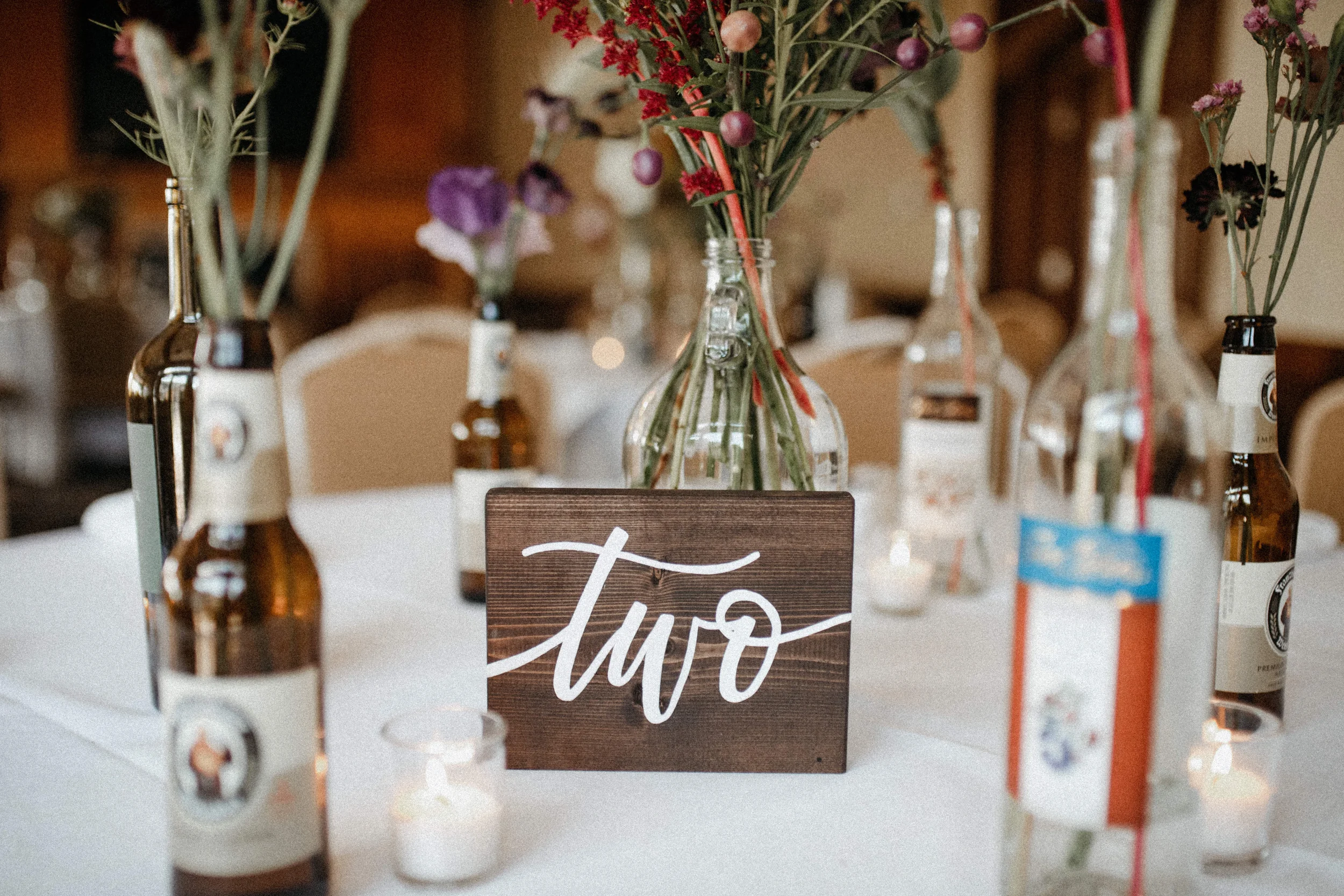 Add warm rustic charm to your reception with a freestanding dark wood block table number. Lettered in bold white cursive script, this sign pairs well with vintage bottle centerpieces, craft beer details, and cozy, casual wedding decor.