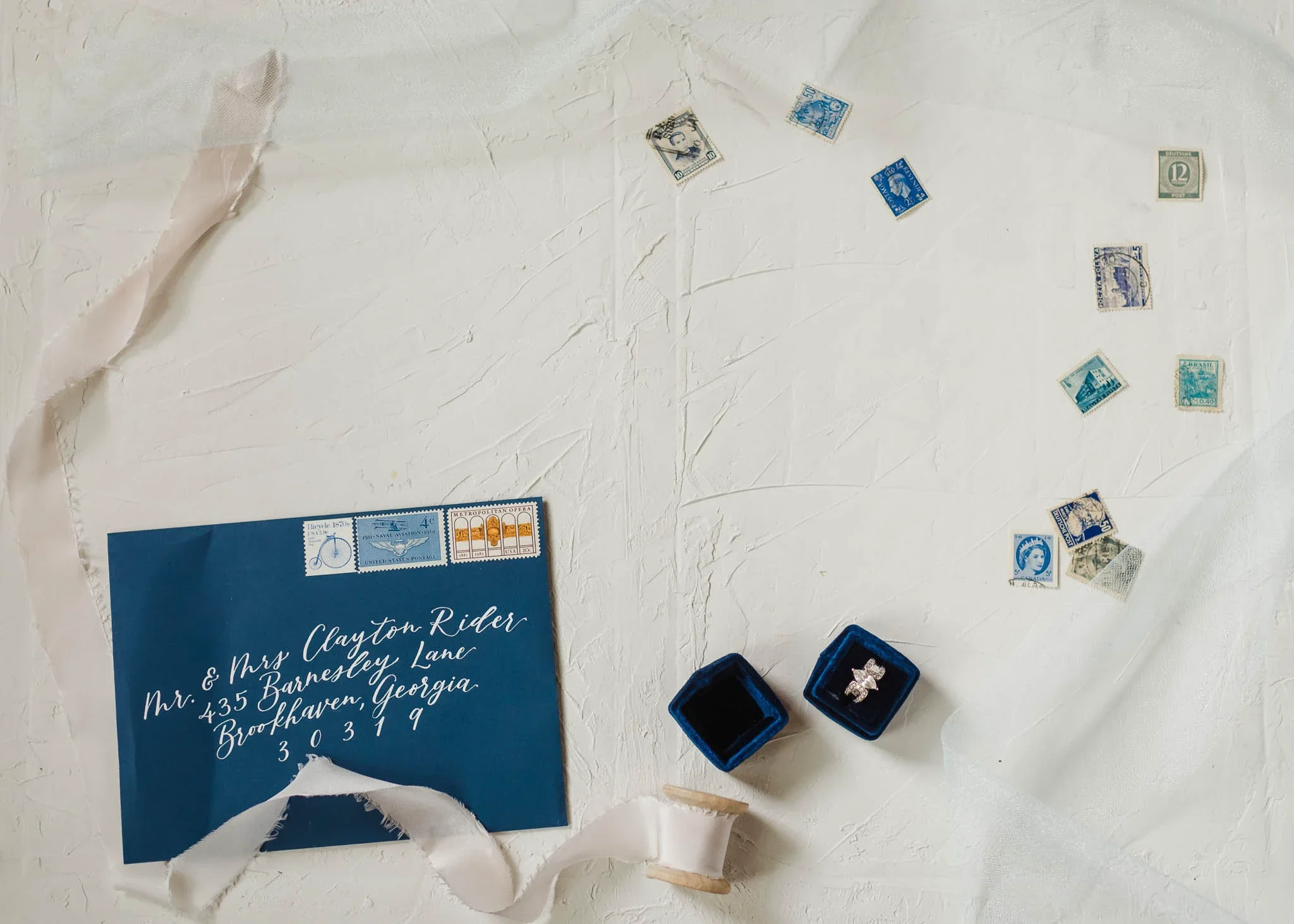 Dark blue navy envelope with white calligraphy address flat lay with navy blue velvet ring box, white silk ribbon, and assorted blue vintage stamps.