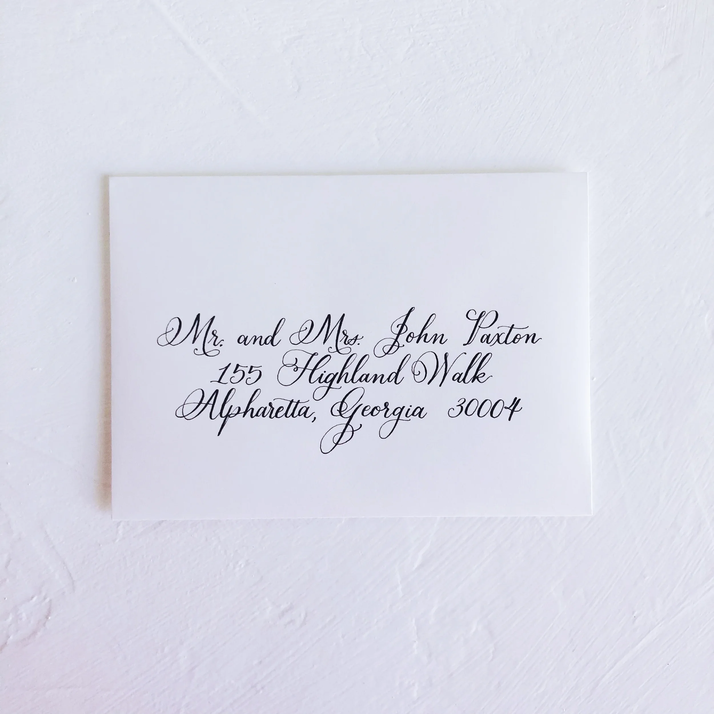 Flat lay of a crisp white wedding invitation envelope addressed in elegant black script calligraphy to Alpharetta, Georgia.