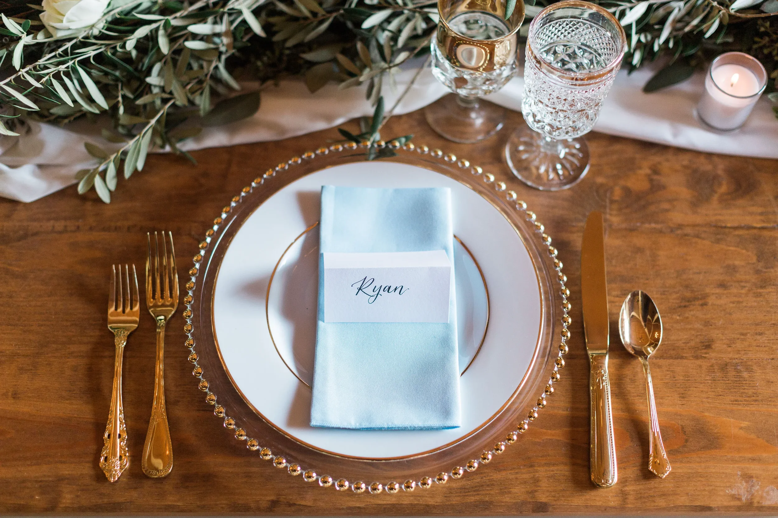 A white tented place card with handwritten script calligraphy reading "Ryan" sits on a light blue napkin, centered on a gold-rimmed white plate and clear beaded charger. The setting rests on a wooden farm table alongside gold flatware, crystal glassw