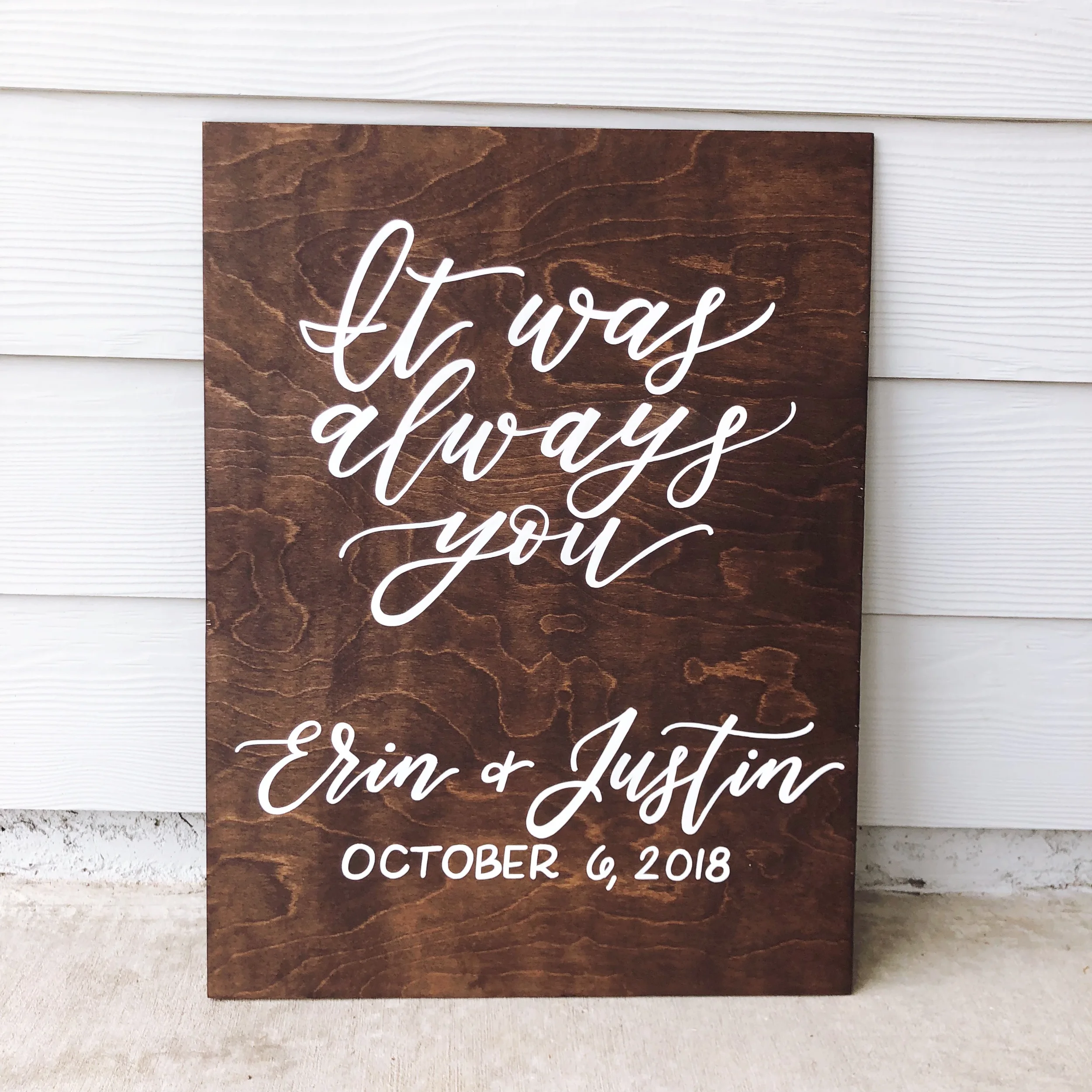 Hand-lettered wooden wedding sign with romantic quote calligraphy set against a white siding backdrop.