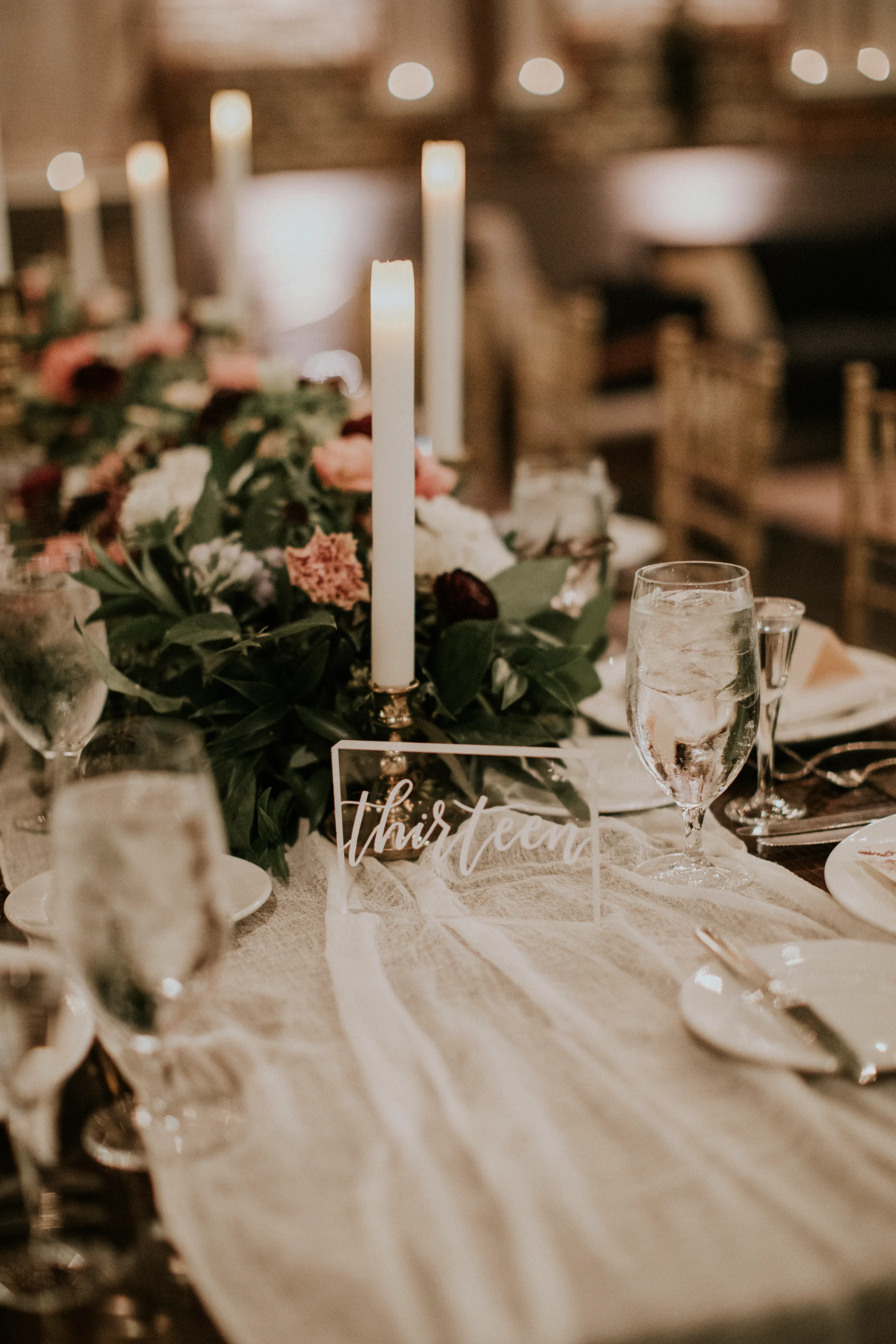 Clear acrylic block wedding table number with white script calligraphy reading "thirteen" placed on a wooden farm table next to a low floral centerpiece, white gauze runner, and tall taper candles.