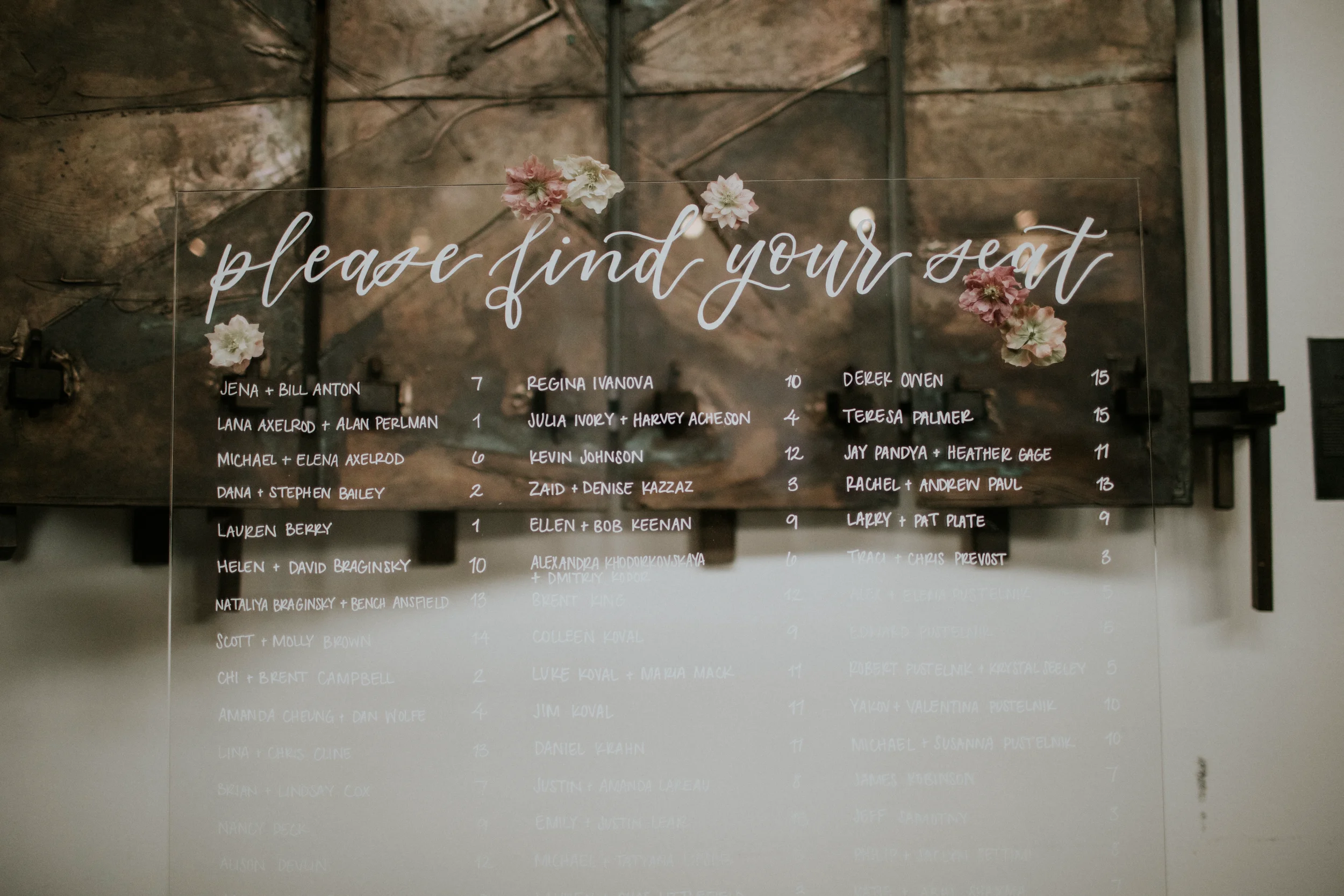Browse our custom acrylic wedding seating chart designed for a modern industrial reception. This large, transparent lucite guest chart features elegant white script calligraphy reading "please find your seat," accented with delicate floral illustrati