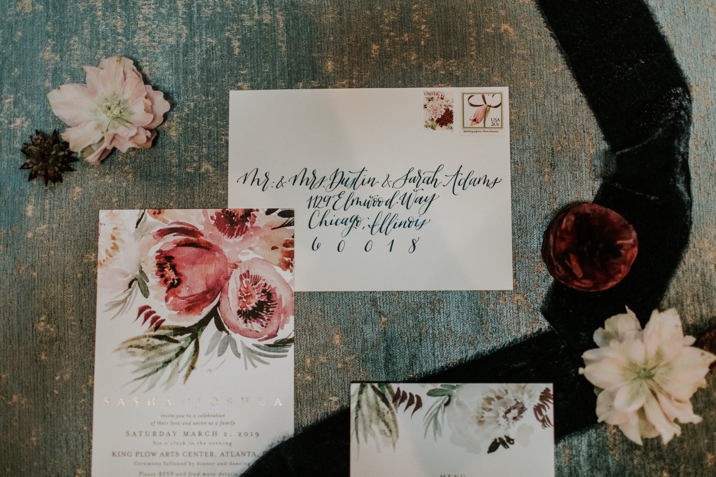 Flat lay overview of a romantic moody hand-addressed calligraphy envelope and a floral watercolor wedding invitation arranged on a textured teal backdrop with a navy velvet ribbon and loose flowers.