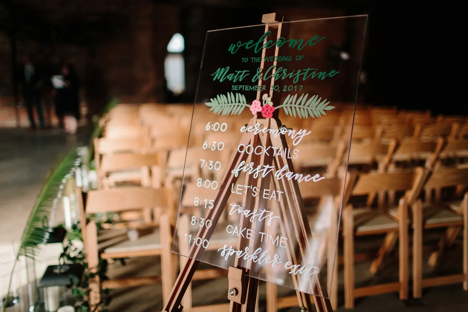 Clear acrylic wedding timeline and welcome sign on a wooden easel with white and green calligraphy.