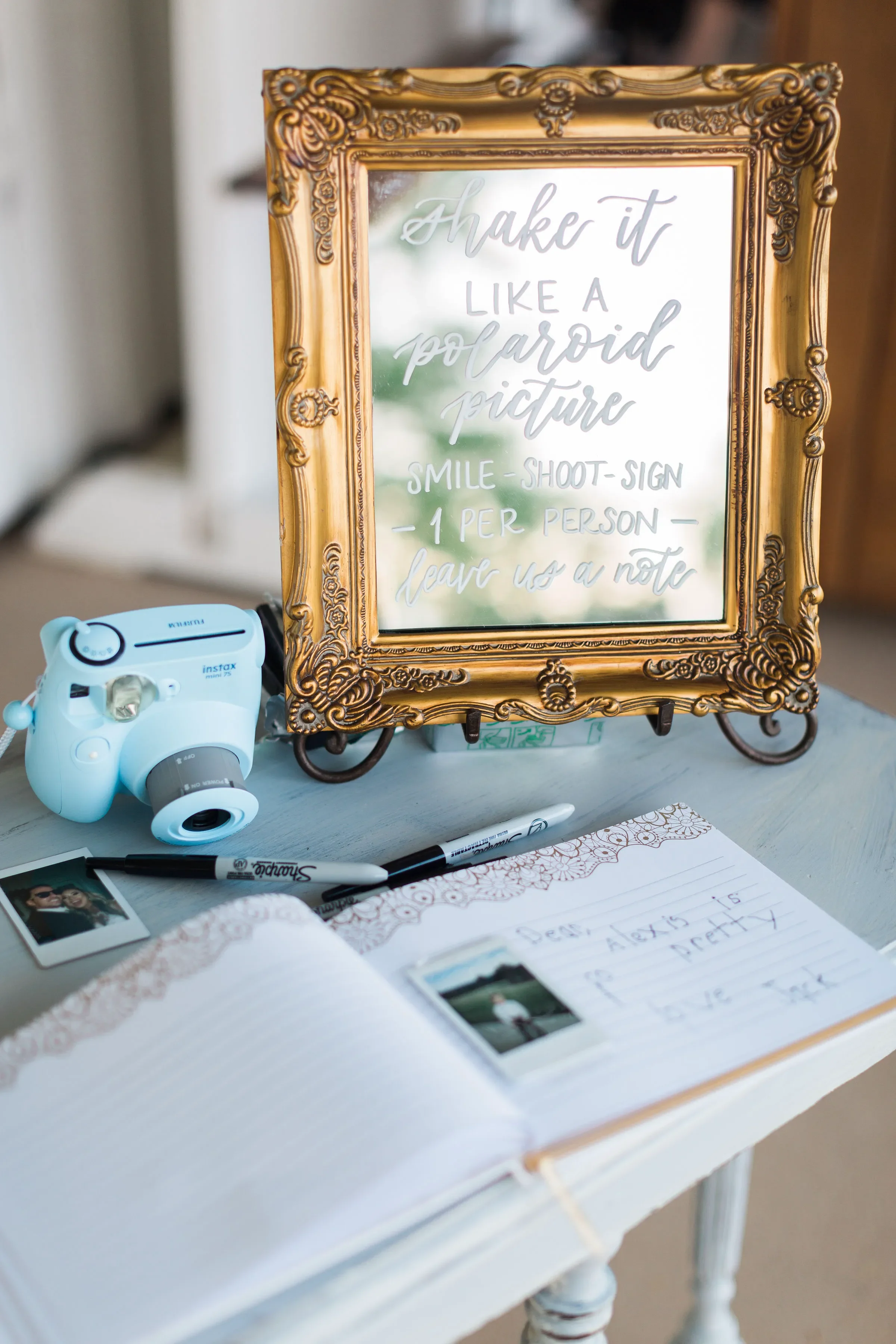 Create a fun, interactive reception station with a custom mirror polaroid guestbook sign featuring hand-lettered white script calligraphy. This ornate, vintage gold-framed mirror displays instructional text reading "Shake it like a Polaroid picture: 