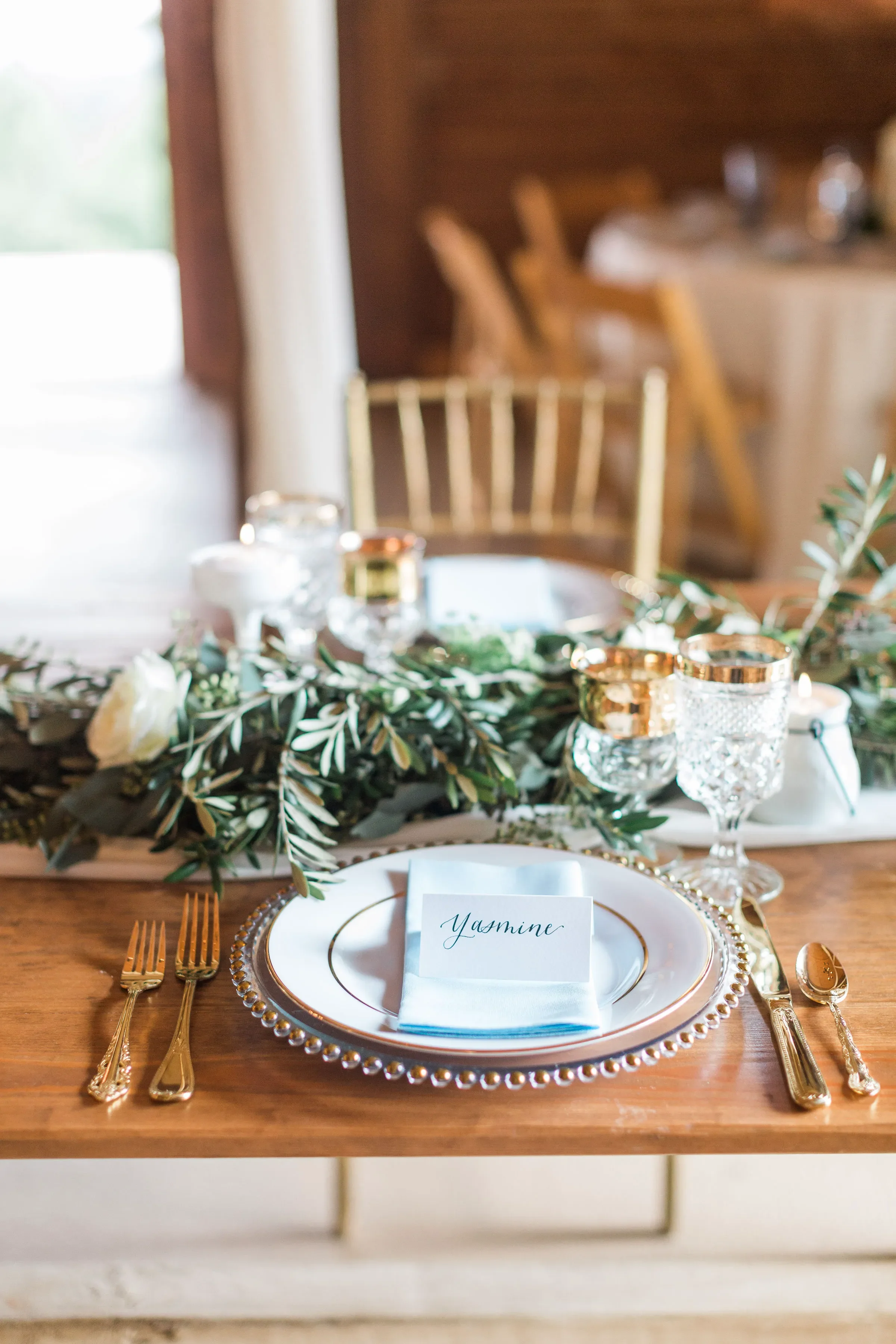 A white tented place card with handwritten script calligraphy reading "Yasmine" sits on a light blue napkin, centered on a gold-rimmed white plate and clear beaded charger. The setting rests on a wooden farm table alongside gold flatware, crystal gla