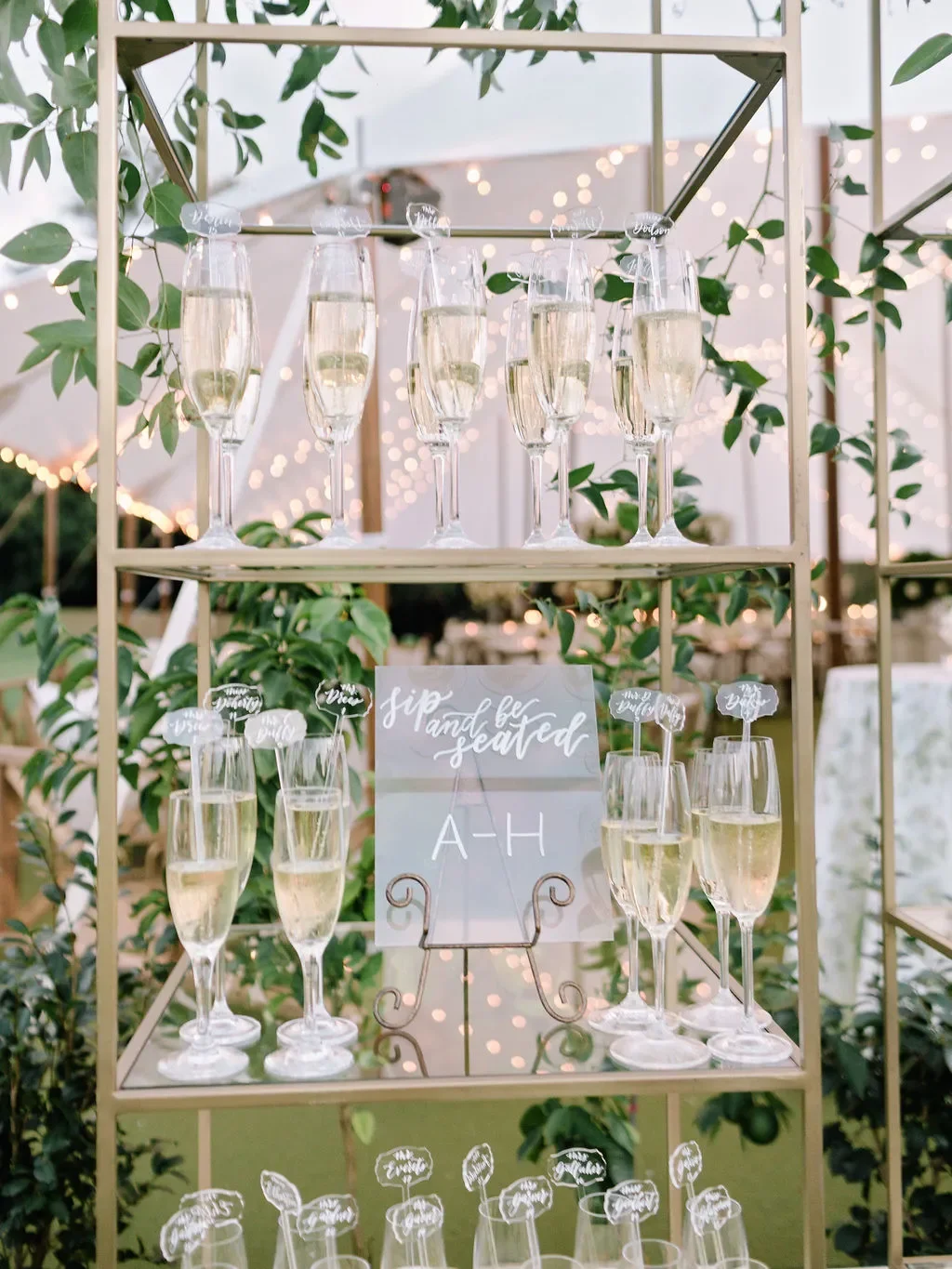 A stunning champagne wall escort card display at an outdoor tented wedding reception. The shelving structure lined with dozens of pre-poured champagne flutes. Lush arrangements of white garden roses, green hydrangea, and trailing eucalyptus cascade a