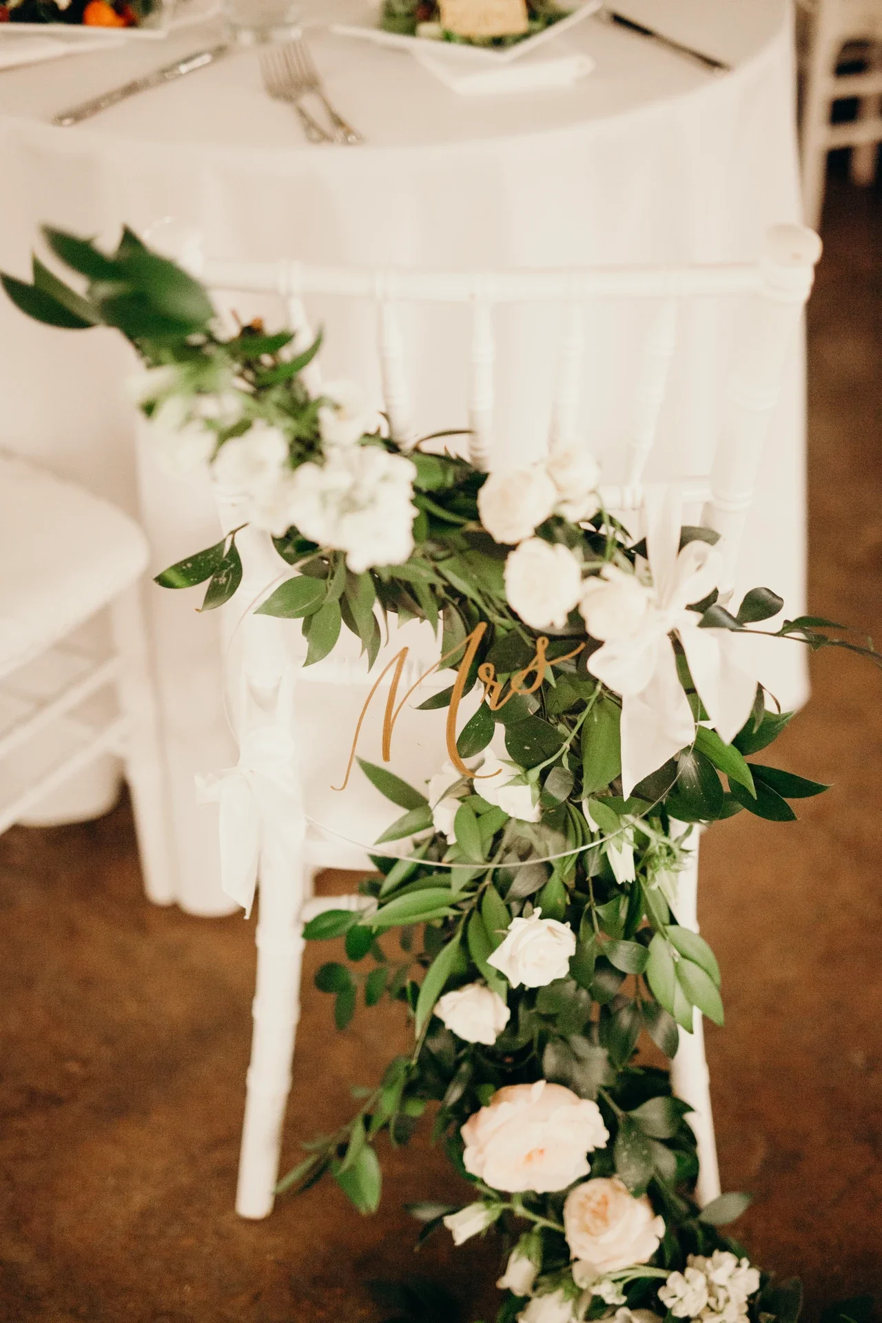 White wedding chair adorned with a sweeping green foliage garland, white roses, and a gold script clear acrylic sign.