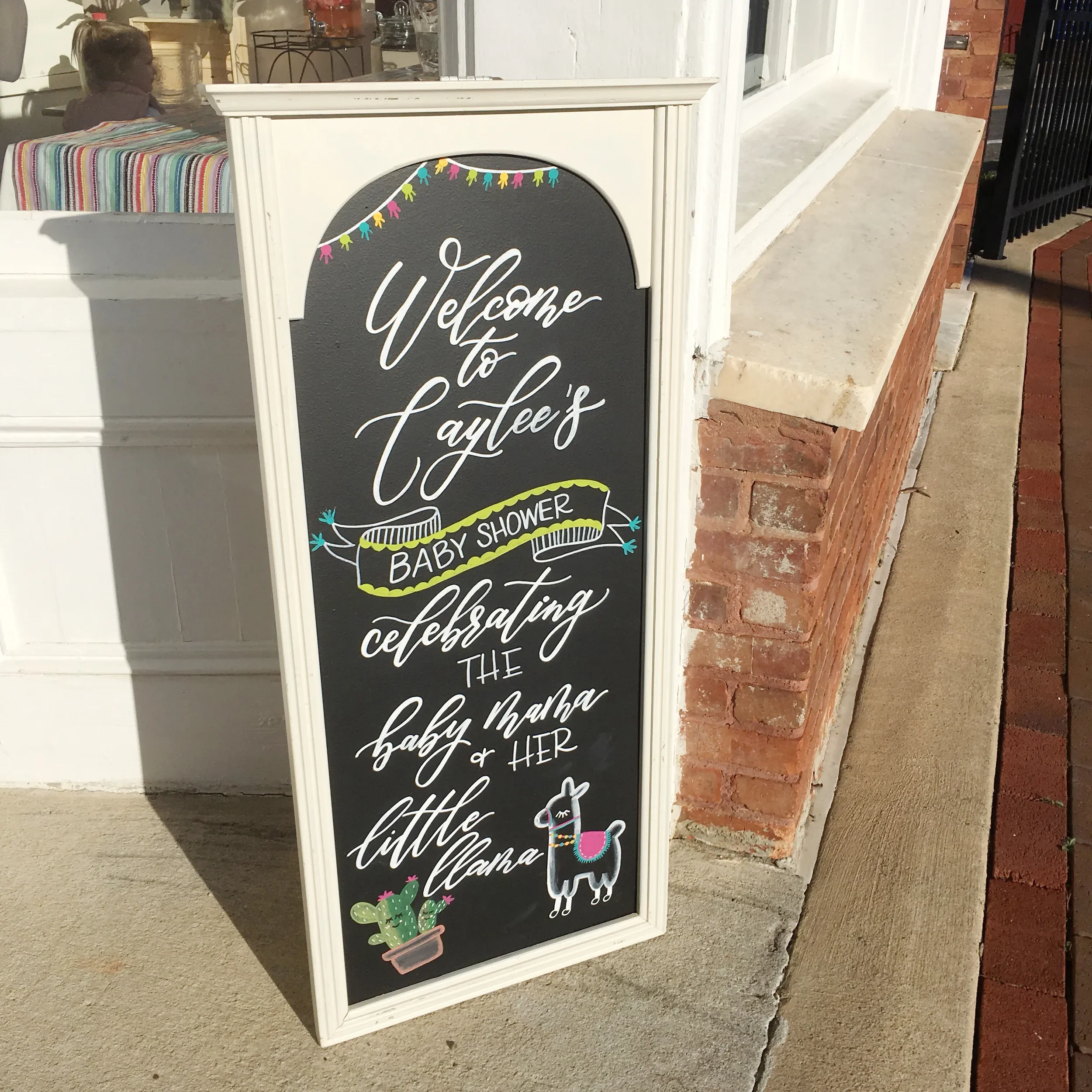 Tall white framed chalkboard welcome sign for a llama-themed baby shower on a sidewalk.