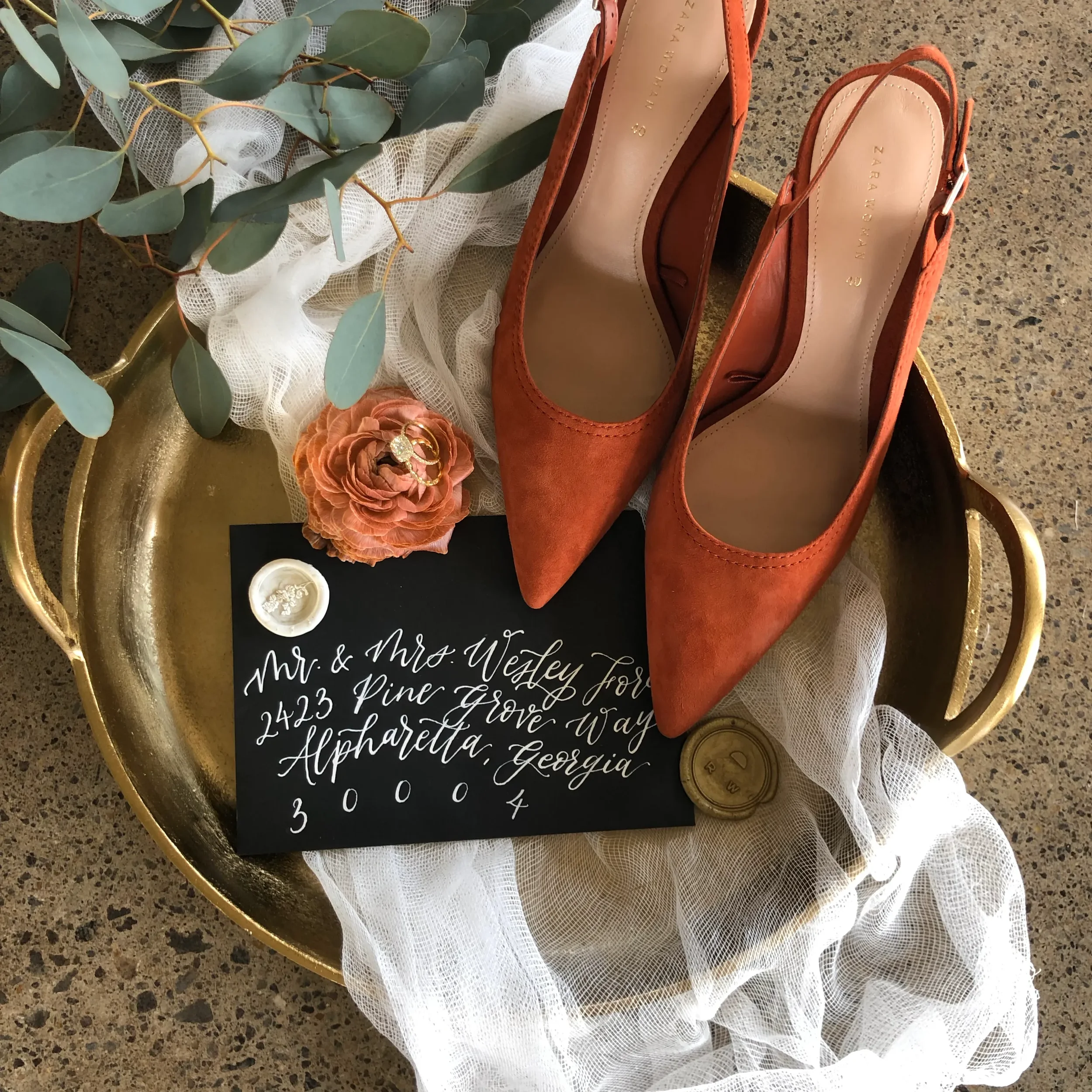 Burnt orange terracotta suede heels styled on a brass tray with a wax-sealed black calligraphy envelope.