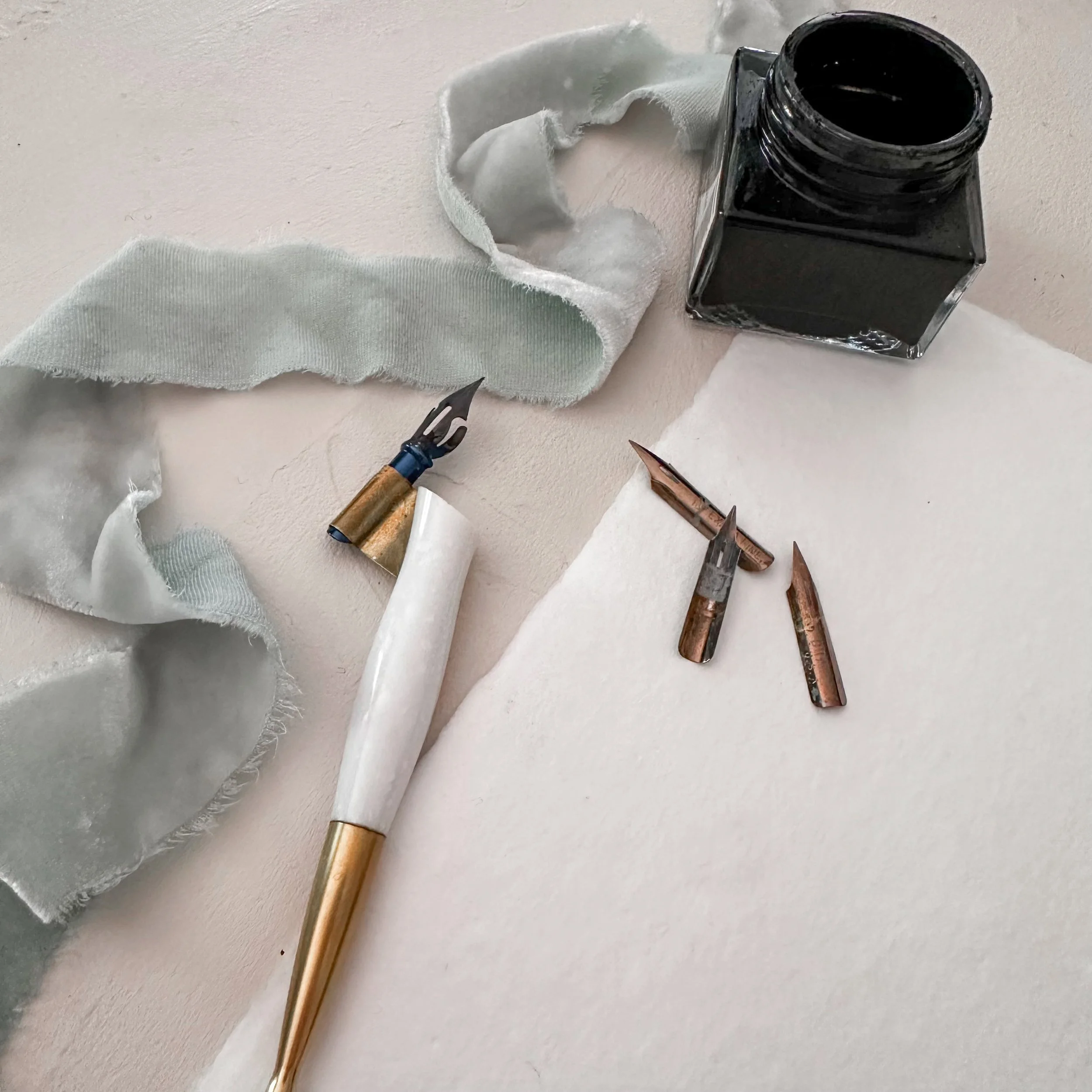 Calligraphy pen, vintage nibs, black ink bottle, and silk ribbon styled on handmade paper.