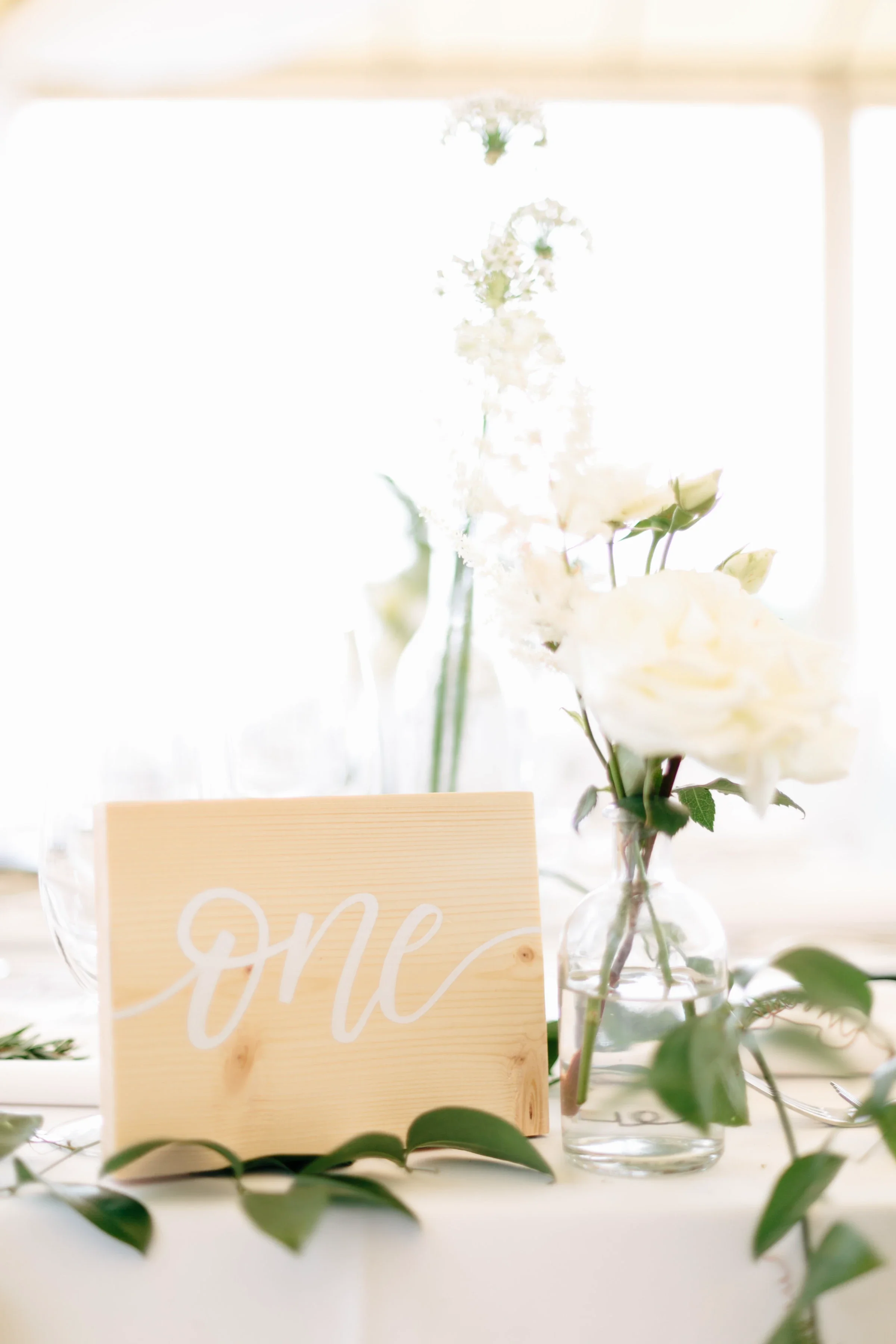 Rustic wood block table number one with white cursive script set amongst greenery and white flowers.
