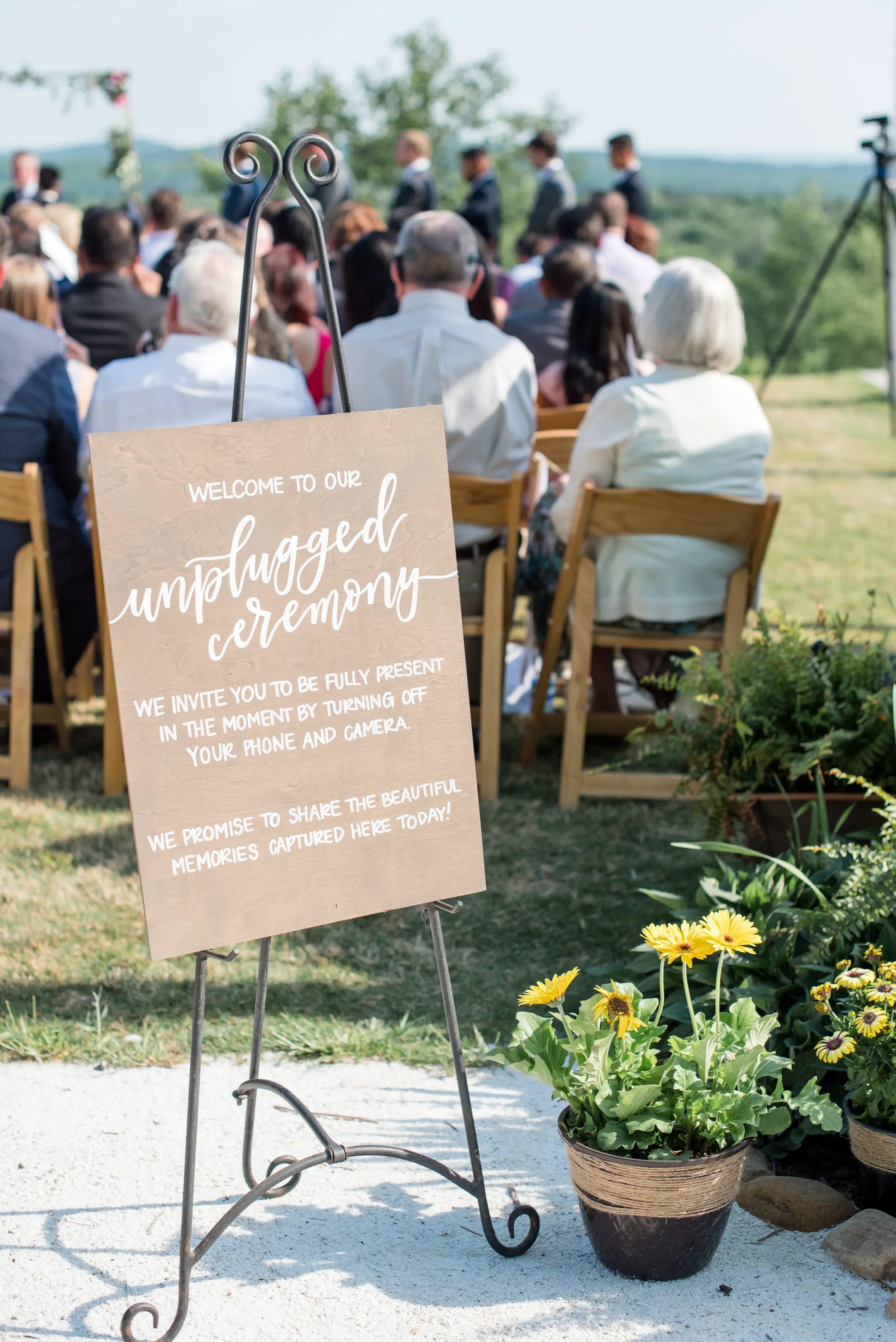 Welcome your guests and set the tone for an intimate, present ceremony with a custom hand-lettered wooden unplugged wedding sign. This classic rustic design perfectly complements a scenic outdoor countryside ceremony space.