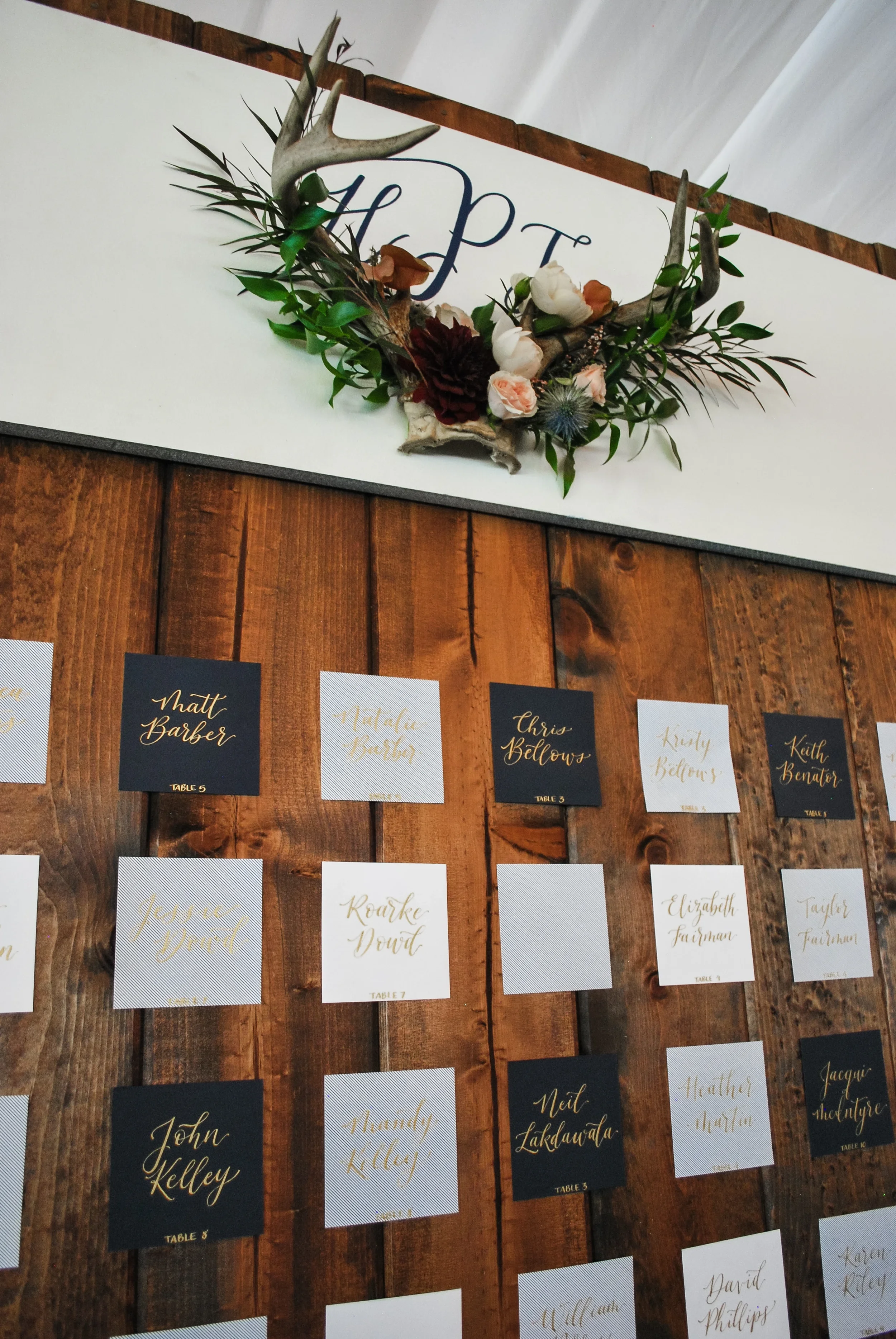 Add rustic warmth to your wedding reception with a custom wood plank escort card wall. Accented by a deer antler crest, natural greenery, and black and slate grey cards lettered in metallic gold calligraphy, this seating display makes a striking stat