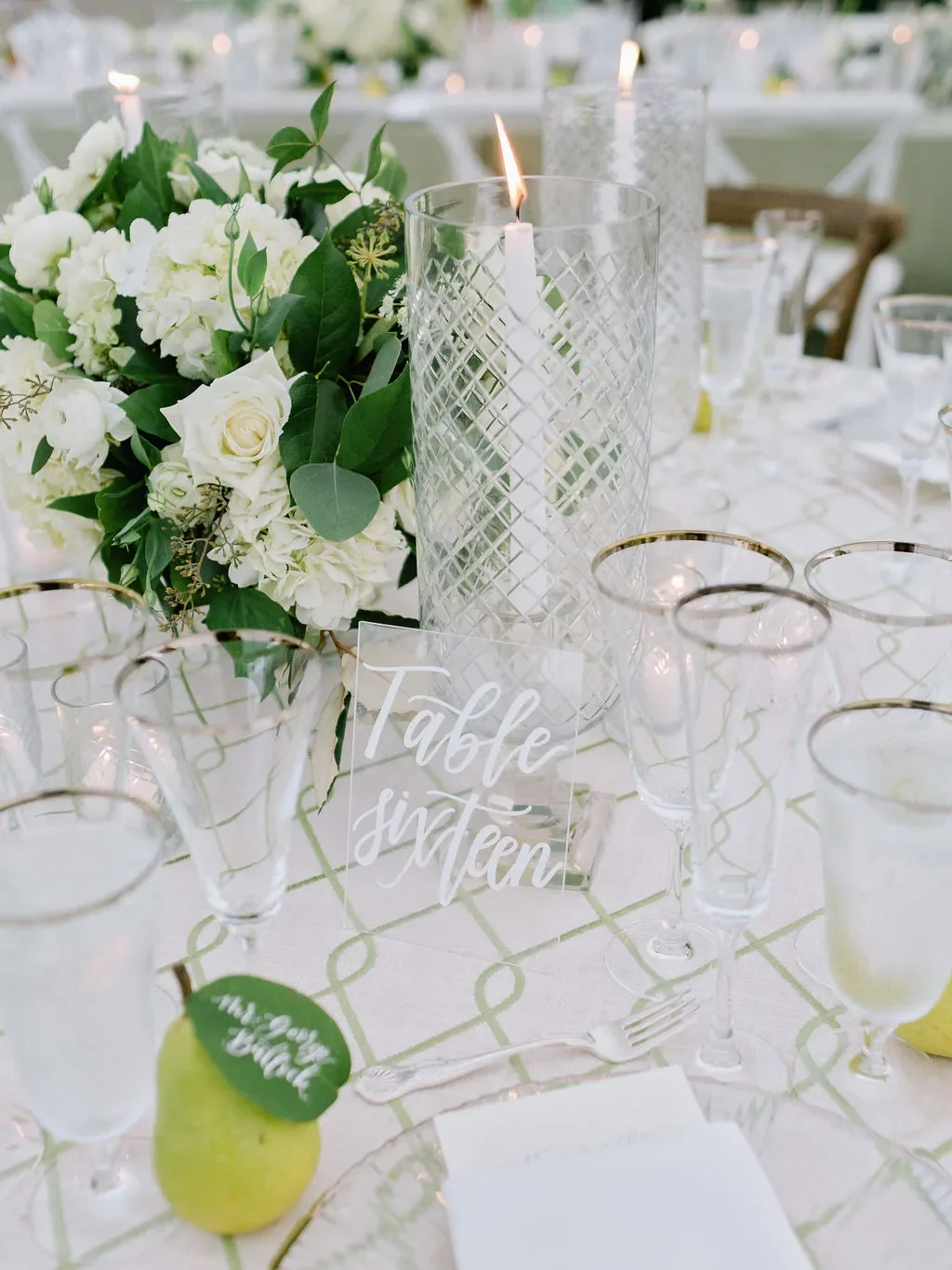 Clear acrylic table number sign with white calligraphy, styled with a hand lettered pear leaf escort card, white floral centerpiece, and elegant wedding reception table decor.
