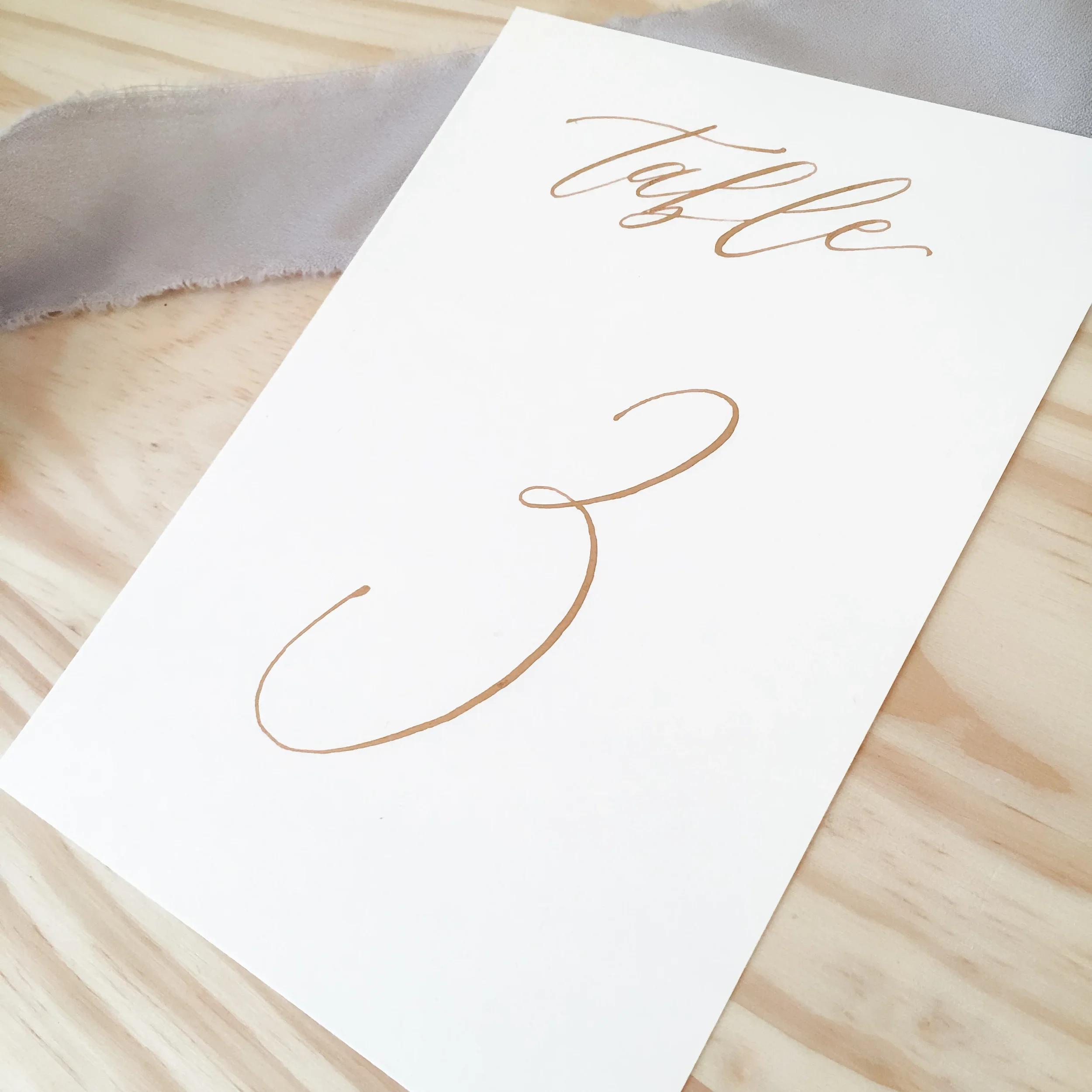 Grid display of classic white paper table number cards with fine-line cursive script in gold calligraphy.