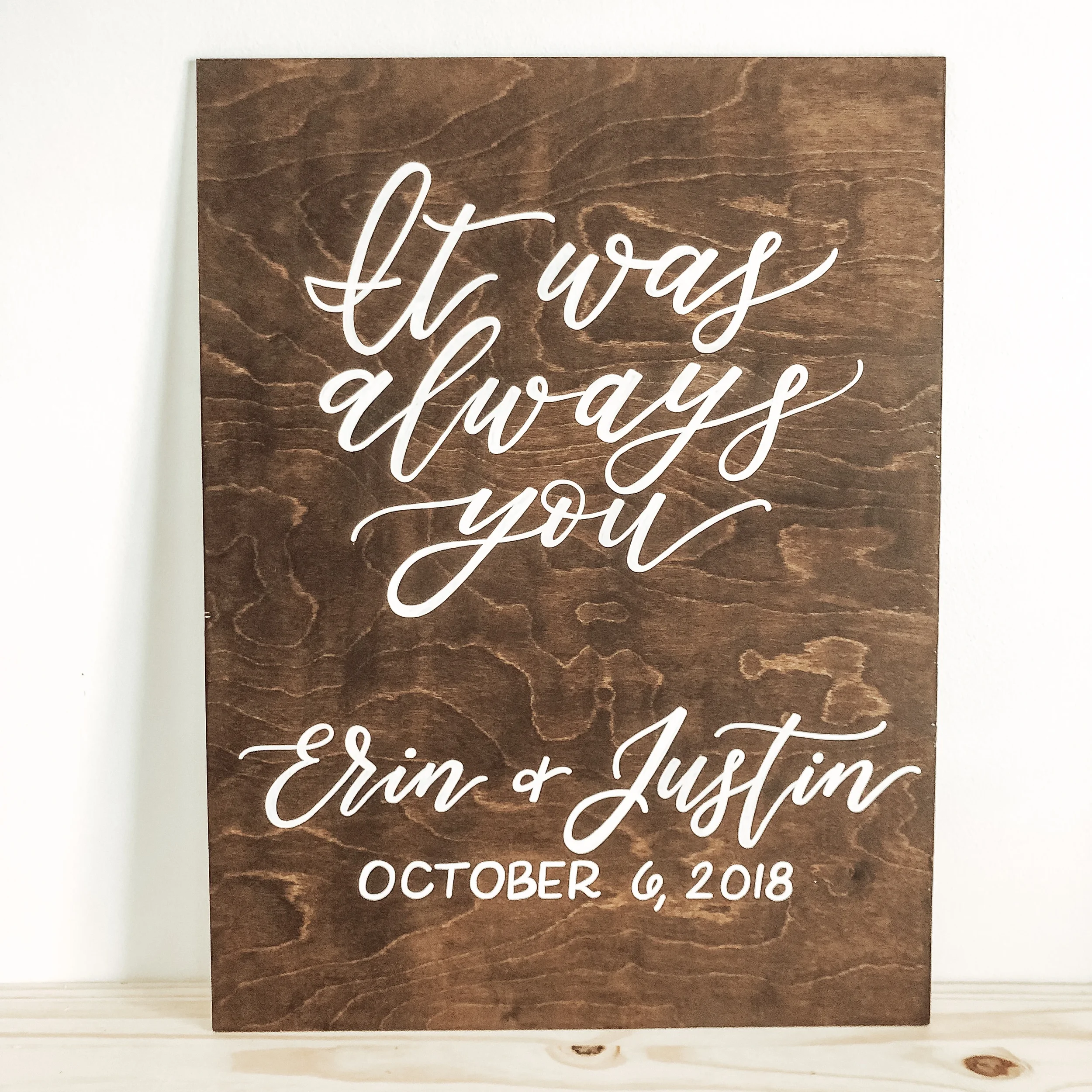 Dark stained wood sign featuring sweeping white script reading "It was always you" with couple's names and date.