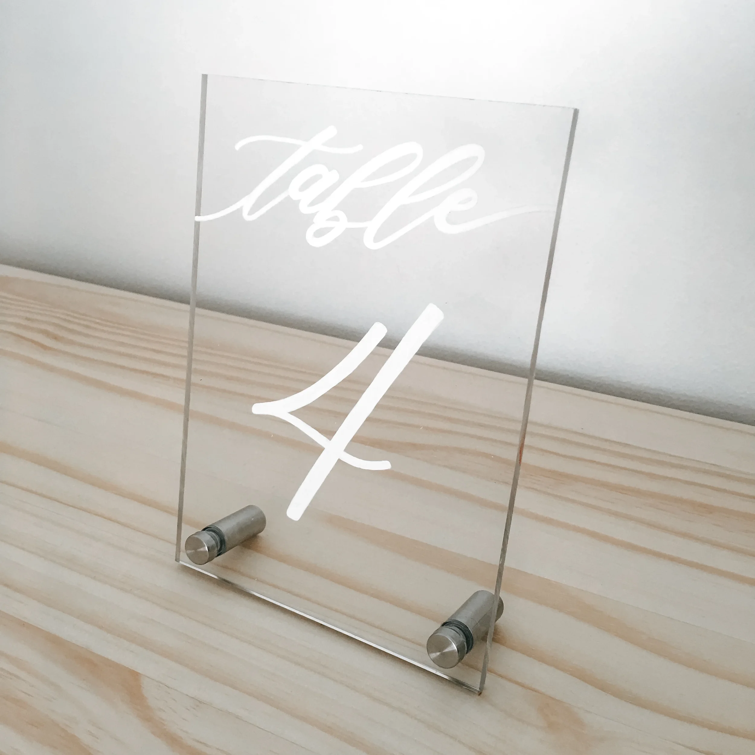 Clear acrylic table number in white script hand-lettering with metal peg stands for a modern style wedding.