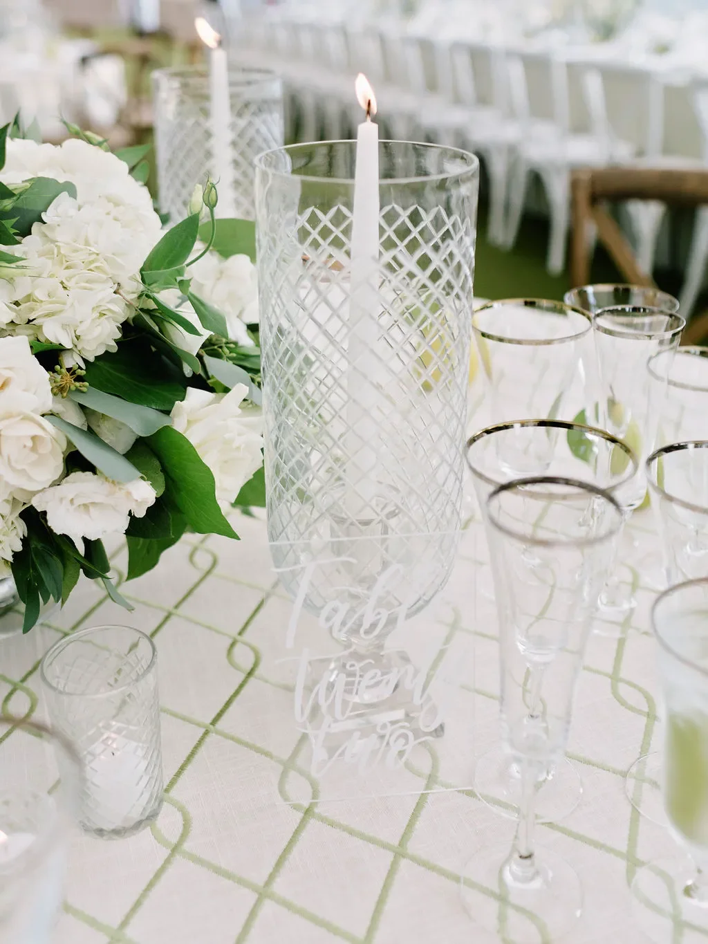 Elegant wedding reception table setting with acrylic table number sign reading "Table Twenty Two," white hydrangea and garden rose centerpiece, silver-rimmed champagne flutes, and white pillar candle in glass hurricane holder.