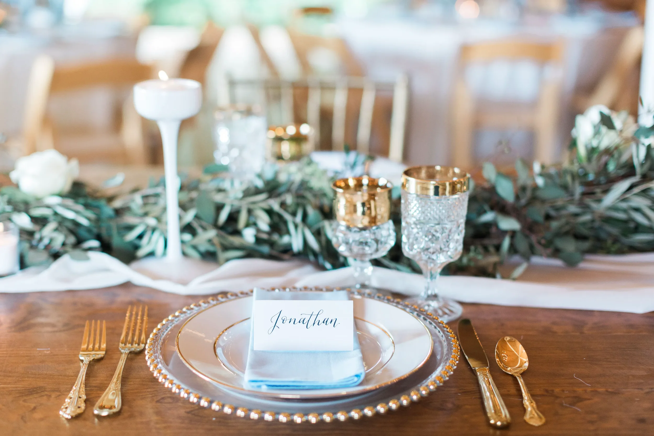 Create a luxurious dining experience with elegant handwritten calligraphy place cards. This custom paper escort card features delicate script lettering, beautifully styled on a soft light blue napkin with gold-rimmed dinnerware and a clear glass bead