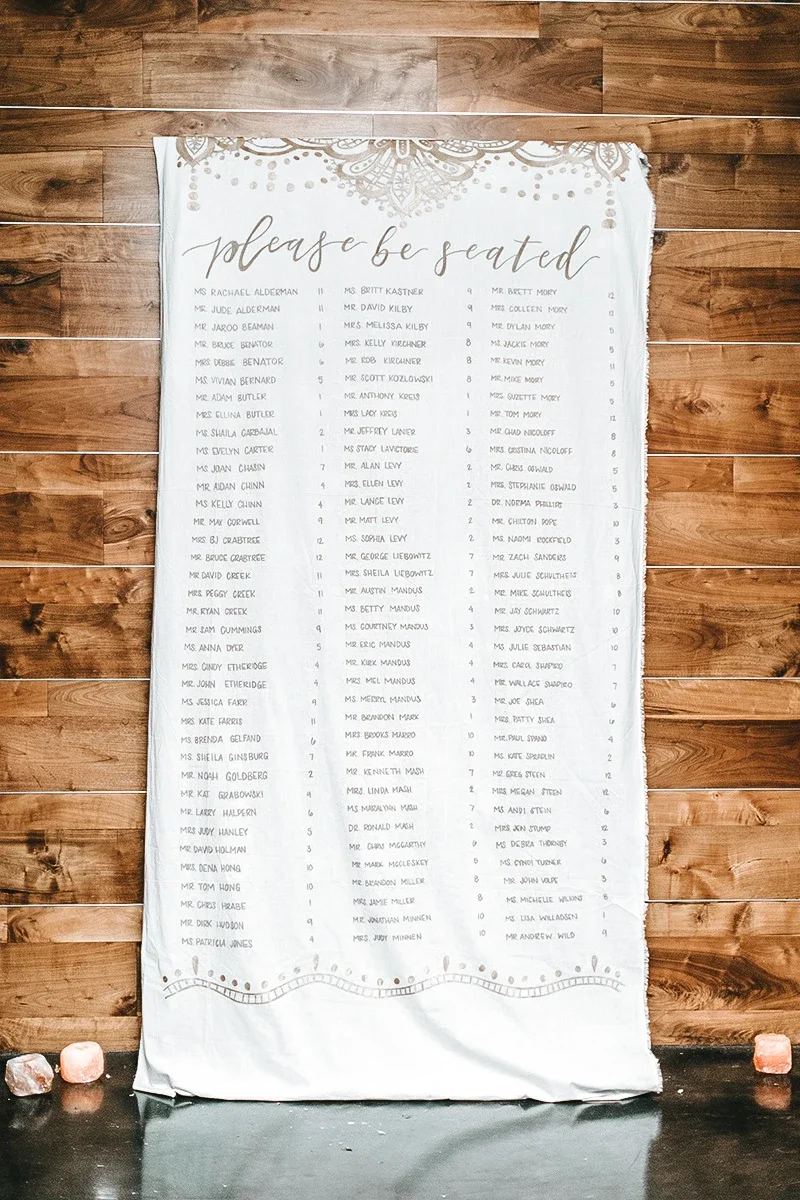 Large white fabric scroll seating chart hanging against a rustic wooden wall reading "please be seated".