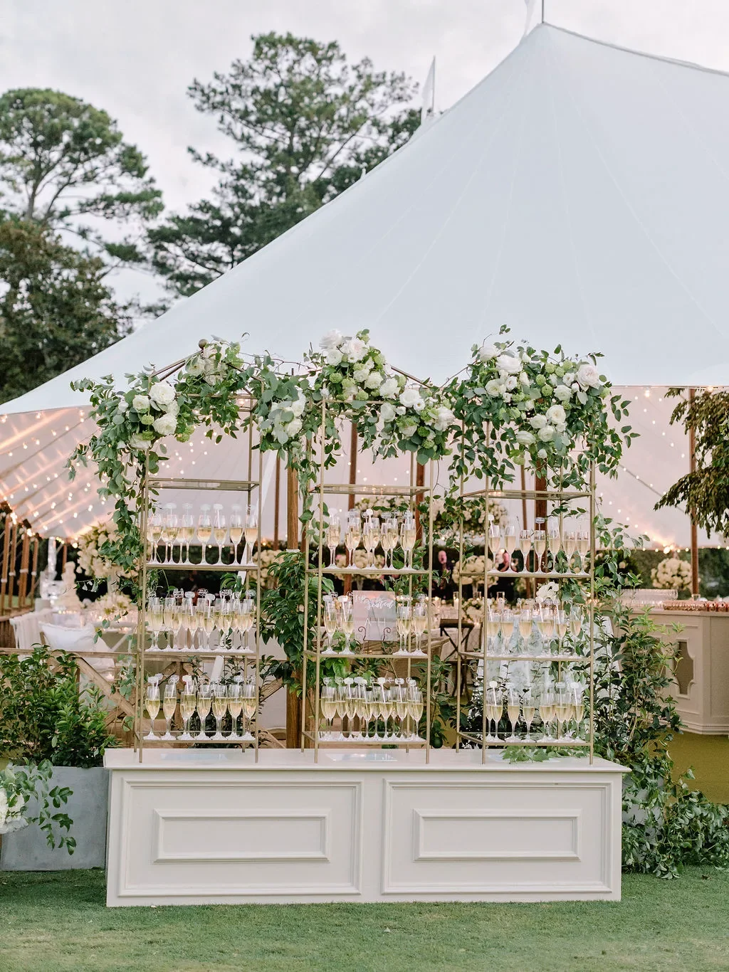 Guests were greeted by this showstopping champagne wall escort card display at the entrance to the tented reception. Hundreds of pre-poured champagne flutes lined gold geometric shelving adorned with cascading white roses, green hydrangea, and eucaly