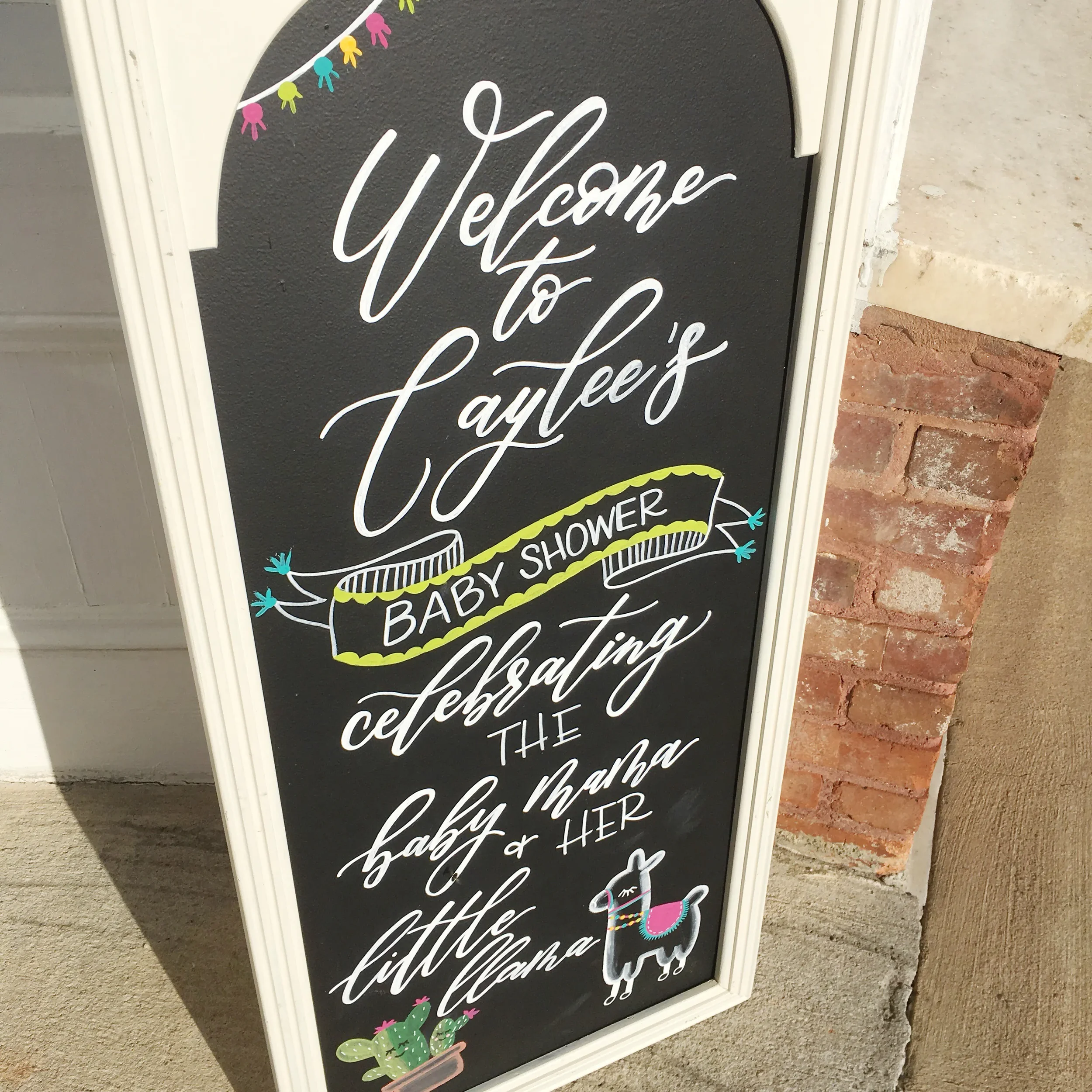 Celebrate the mama-to-be with an eye-catching, custom-designed baby shower chalkboard sign. Featuring a clean white frame, modern script calligraphy, and colorful hand-drawn illustrations of a llama and desert cacti, this welcoming sign adds a bright