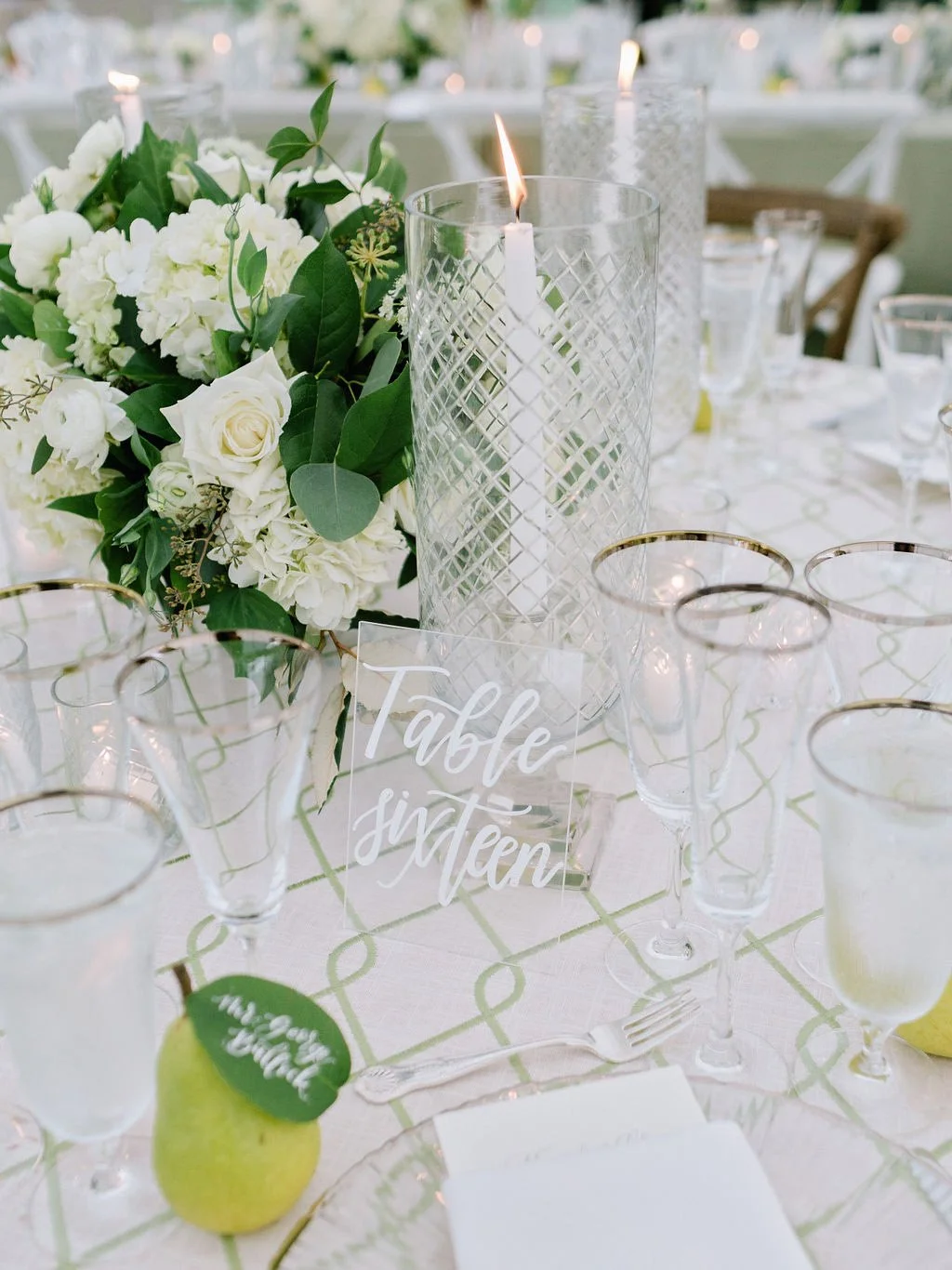 Clear acrylic table number sign with white calligraphy, styled with a hand lettered pear leaf escort card, white floral centerpiece, and elegant wedding reception table decor.