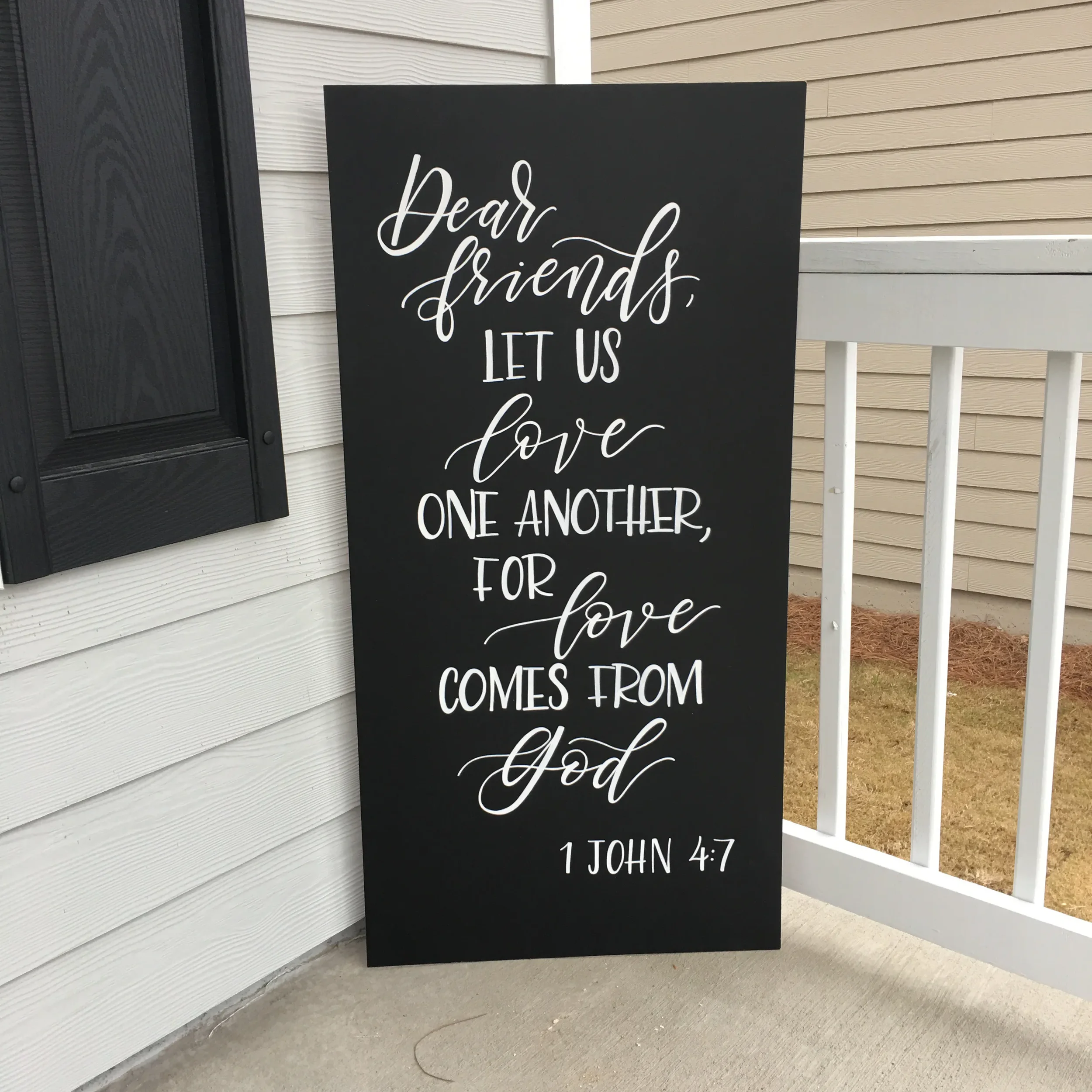 Tall chalkboard wedding sign displaying a hand-lettered 1 John 4:7 Bible verse on a white porch.