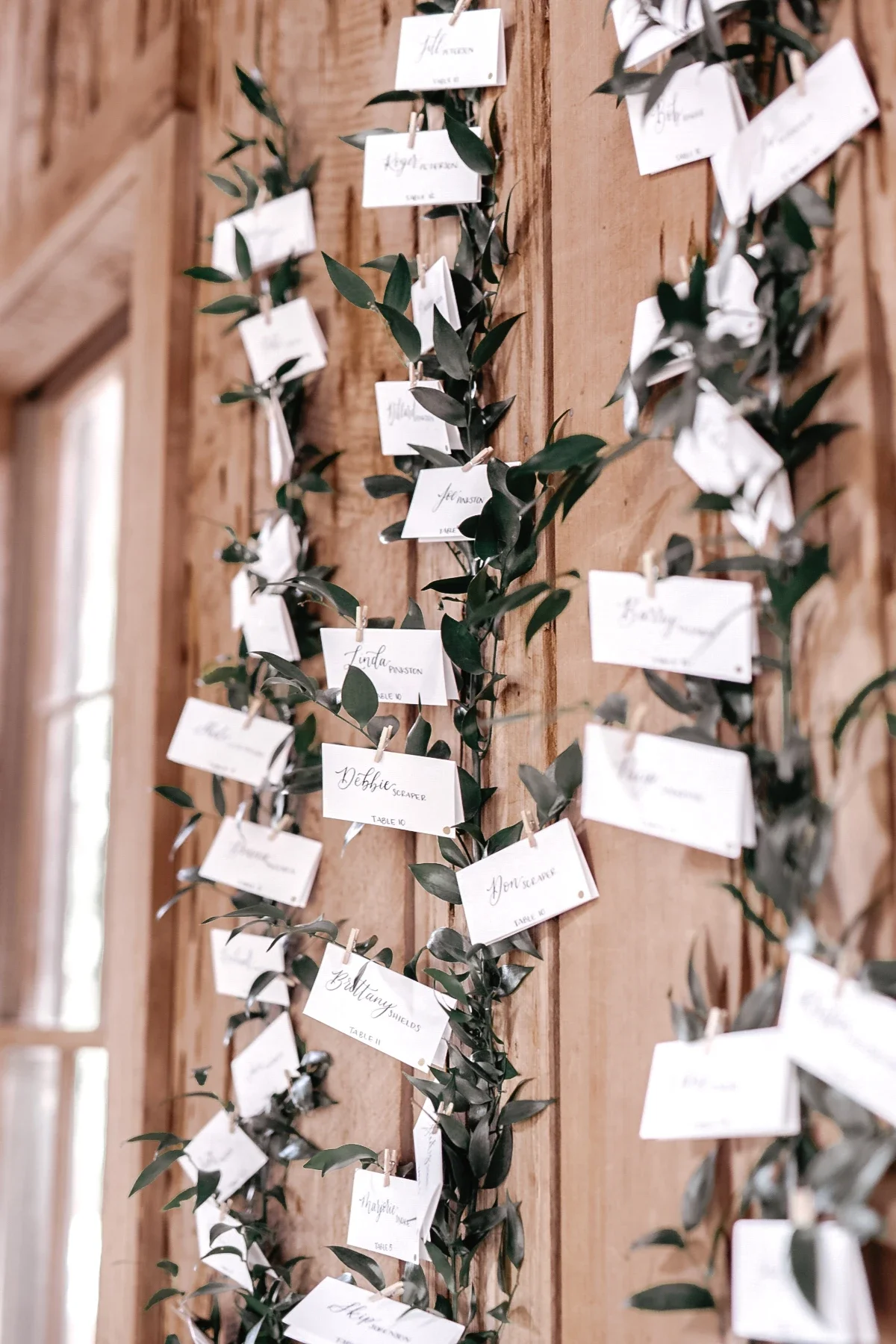Rustic wooden wall wedding seating display featuring calligraphed guest name cards attached to lush leaf garlands.
