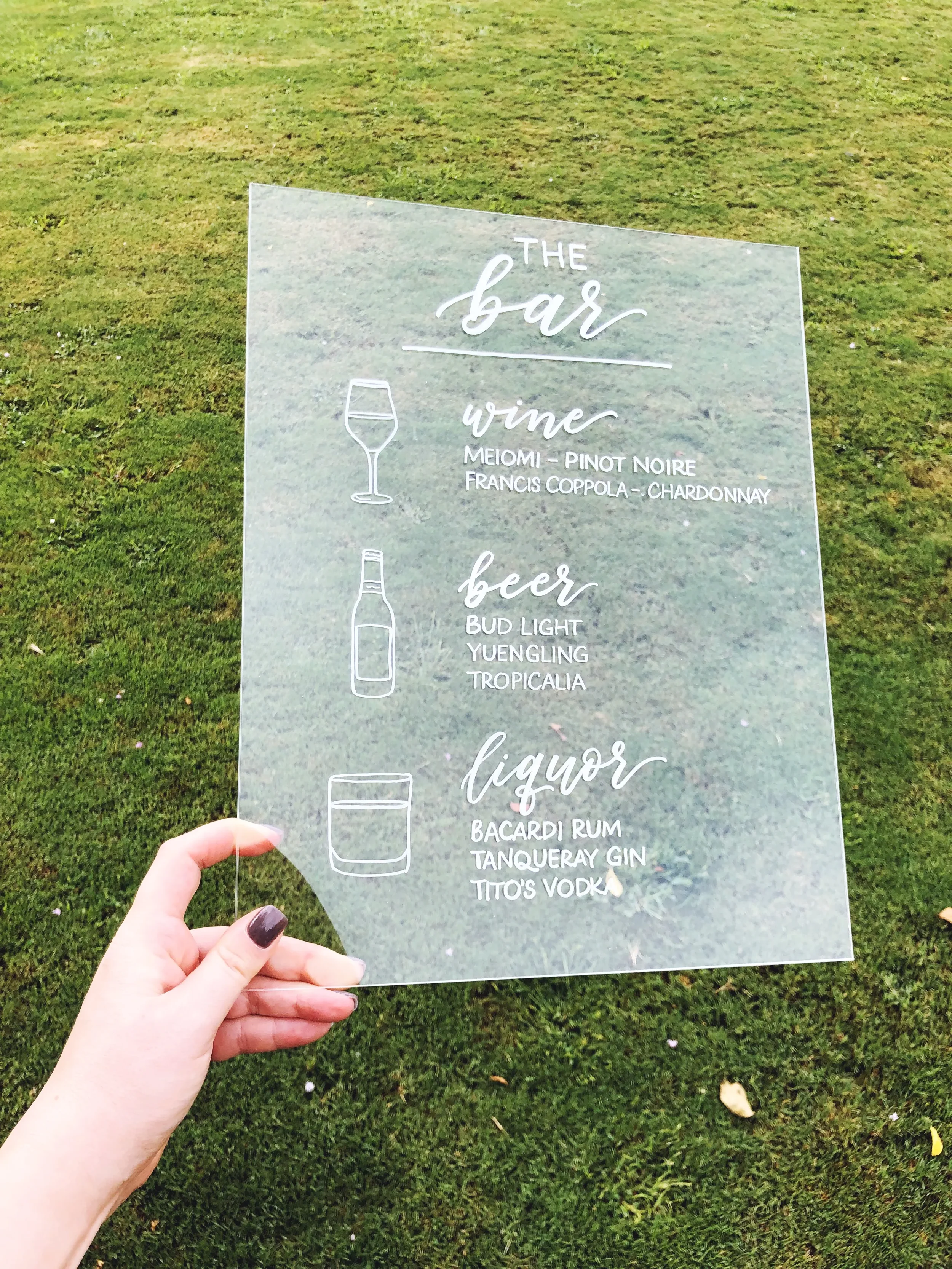 Elevate your cocktail hour setup with a sleek, hand-lettered clear acrylic bar menu sign. Combining modern script typography with clean, white line-art illustrations of glassware, this transparent menu piece layers effortlessly against natural outdoo
