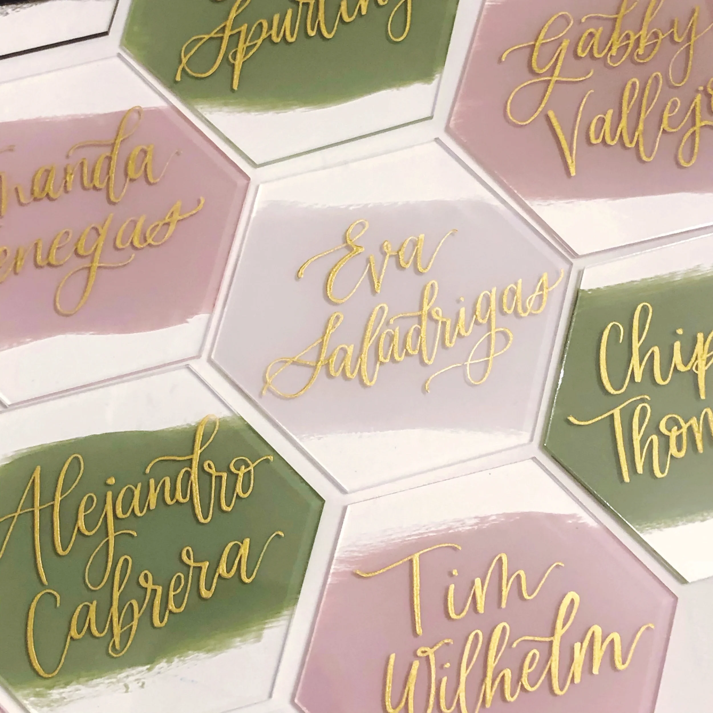 Mosaic array of geometric hexagon place cards with painted backs in sage green, white, and blush pink with gold calligraphy.