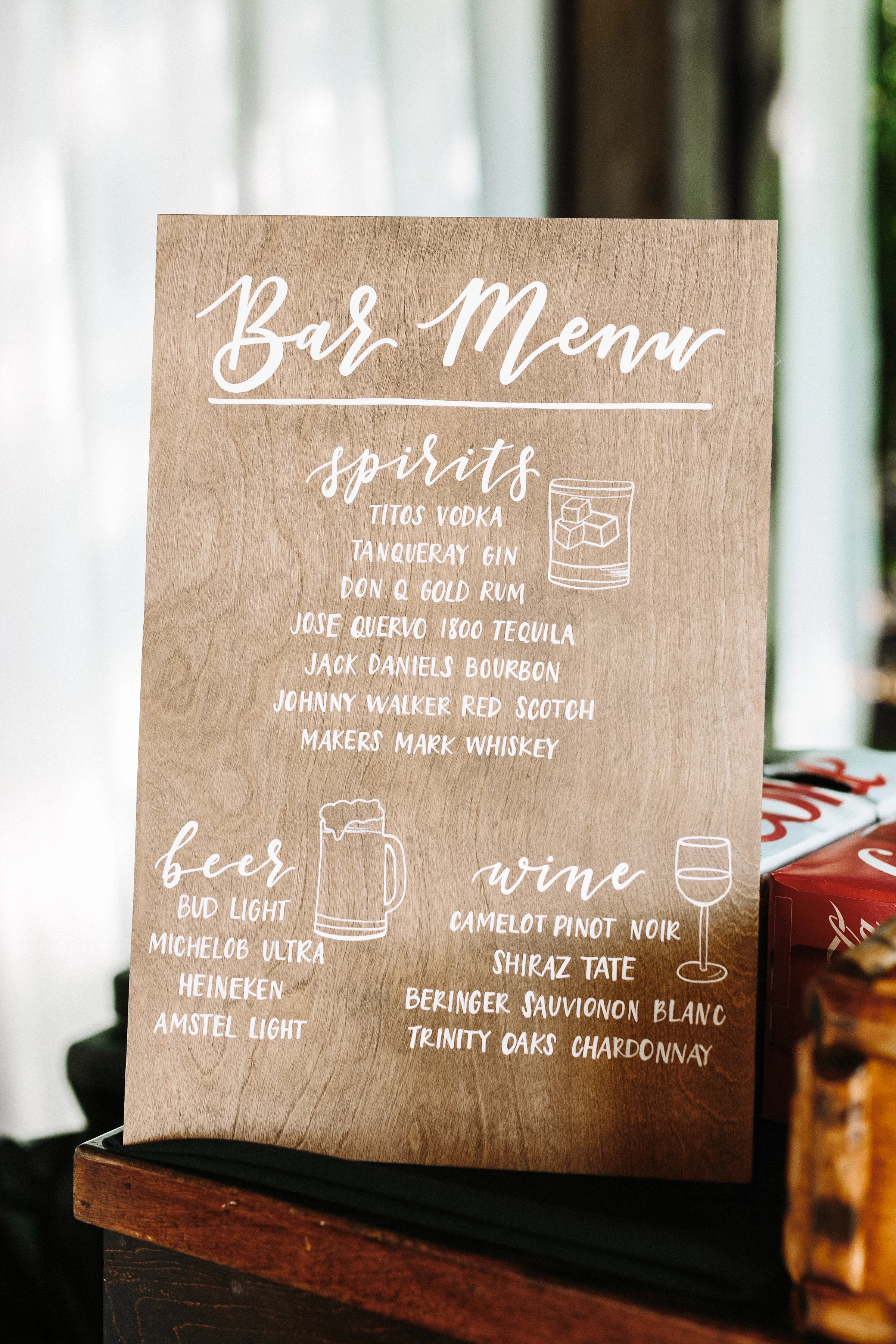 Elevate your beverage station with a custom hand-lettered wooden bar menu sign. Detailed with delicate white typography and custom line drawings of beer mugs and wine glasses, this rustic board keeps bar service clear and visually engaging.