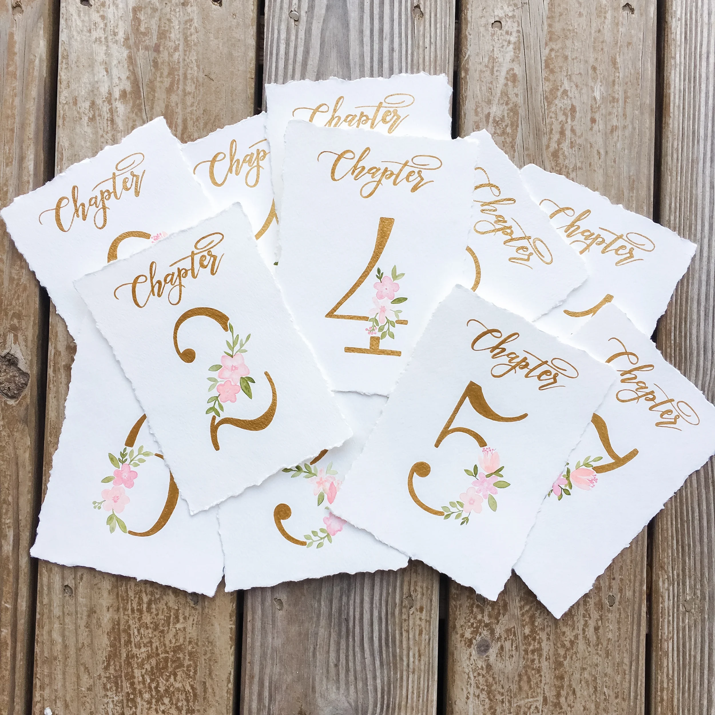 Custom deckled-edge table numbers as "chapters" with gold script and painted pink flowers for a romantic book lover wedding.