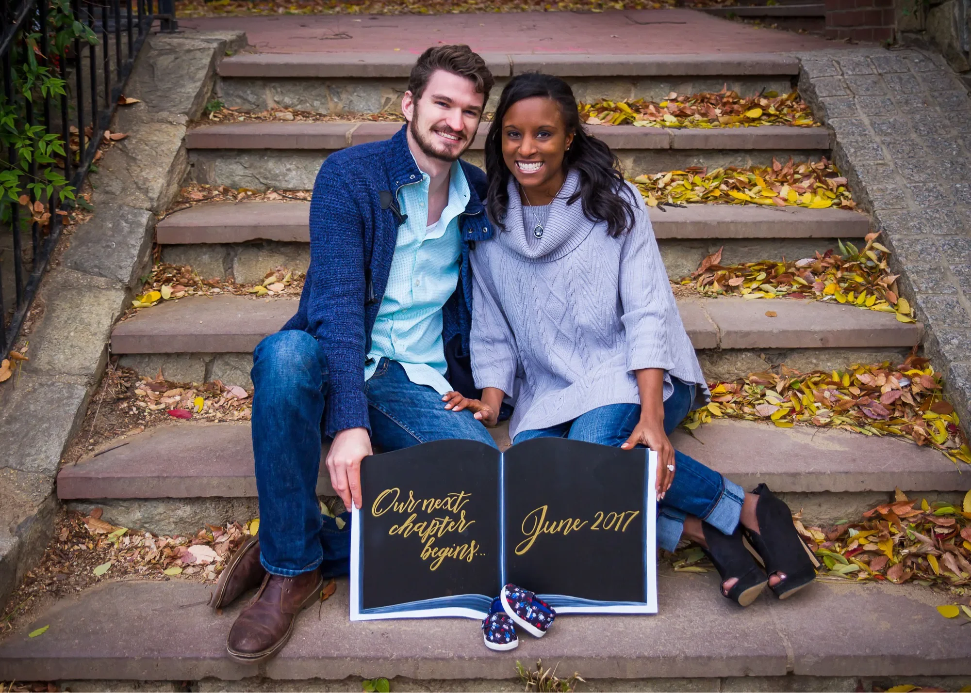 Couple sitting on outdoor steps behind an open-book shaped chalkboard displaying their baby's due date.