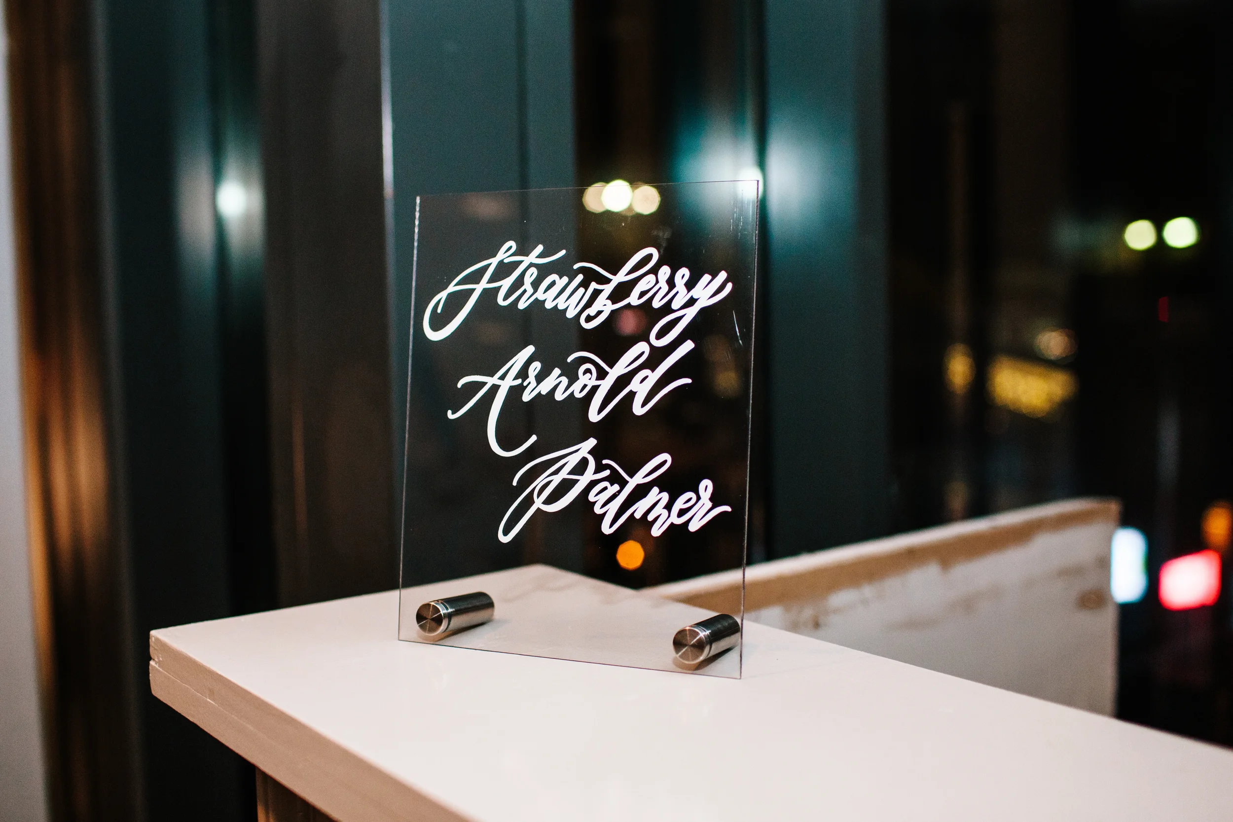 Sleek white ink acrylic bar sign listing custom signature wedding cocktails.