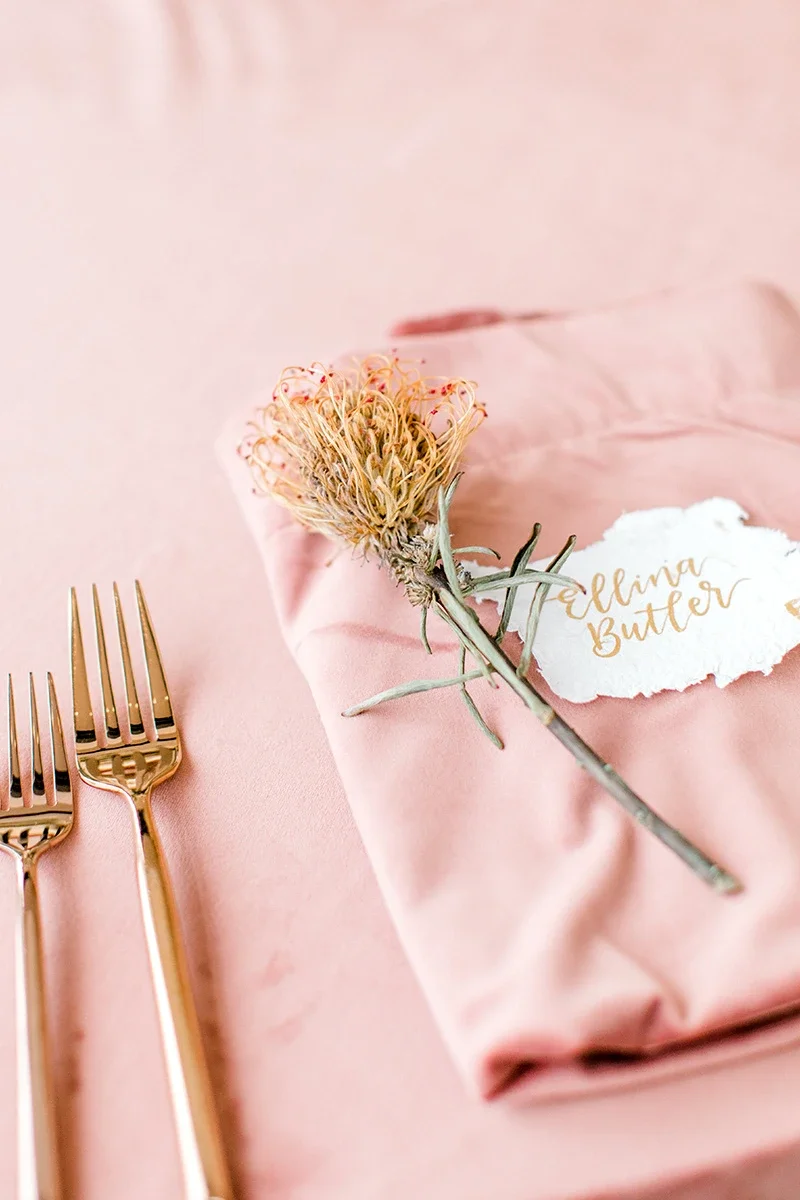 Deckled-edge paper place card written in metallic gold calligraphy script on a folded dusty blush pink napkin with a pincushion protea stem.
